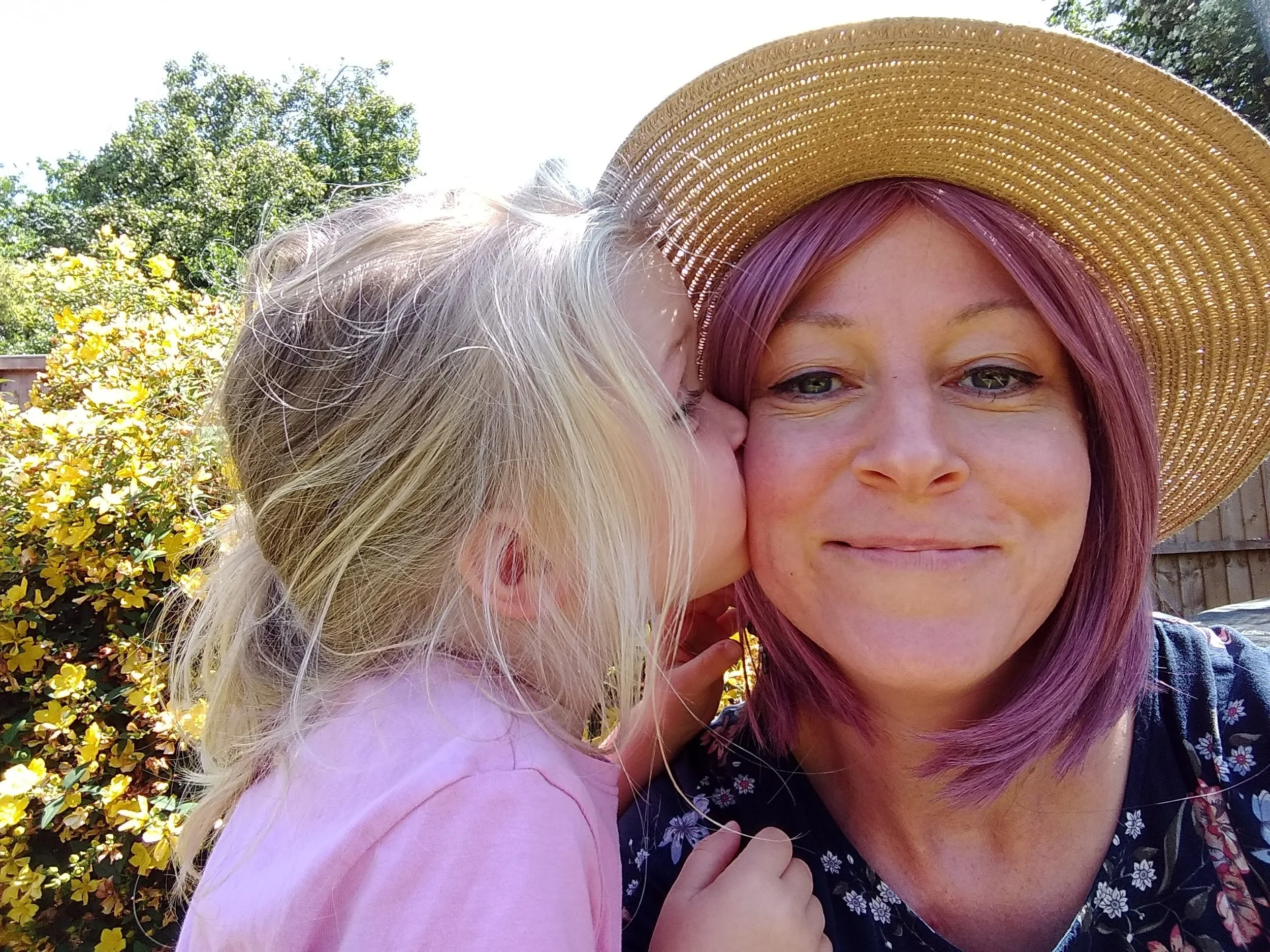 A smiling woman with pink hair wearing a large straw hat, hugging a young girl with blonde hair outdoors, with yellow flowering bushes and green trees in the background.