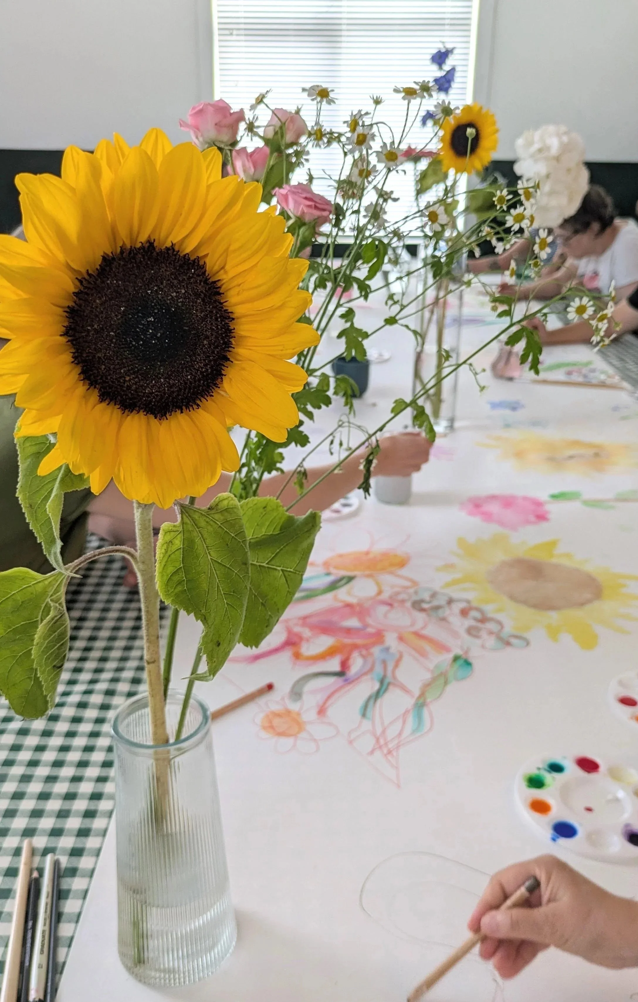 Close-up of a clear ribbed glass vase with a sunflower, pink roses, daisies, and small blue flowers on a table, with people painting large flowers on a mural in the background.