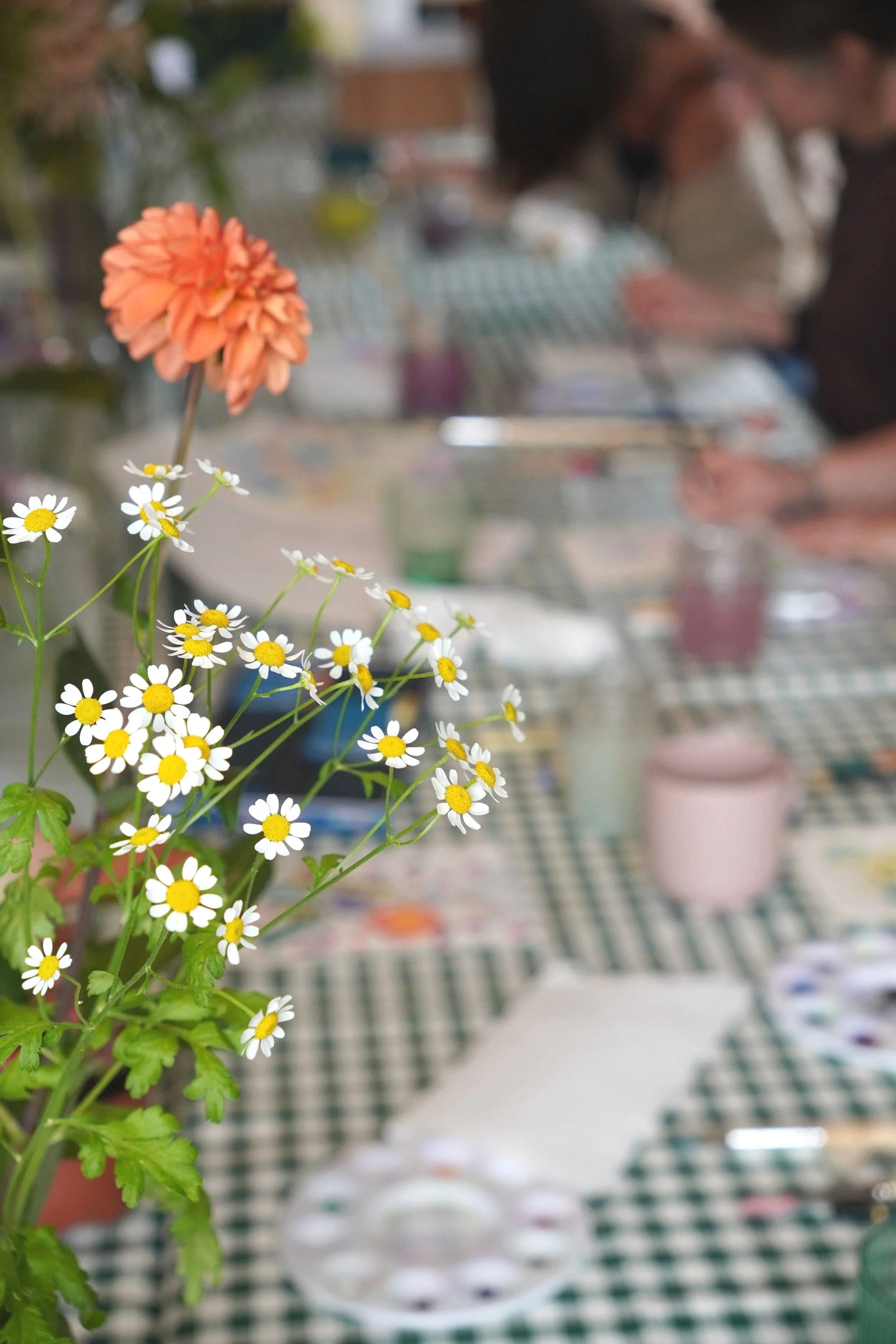 Close-up of white daisies with yellow centers and an orange flower in the background, in front of a blurred indoor scene with people making colourful paintings on a checkered tablecloth.