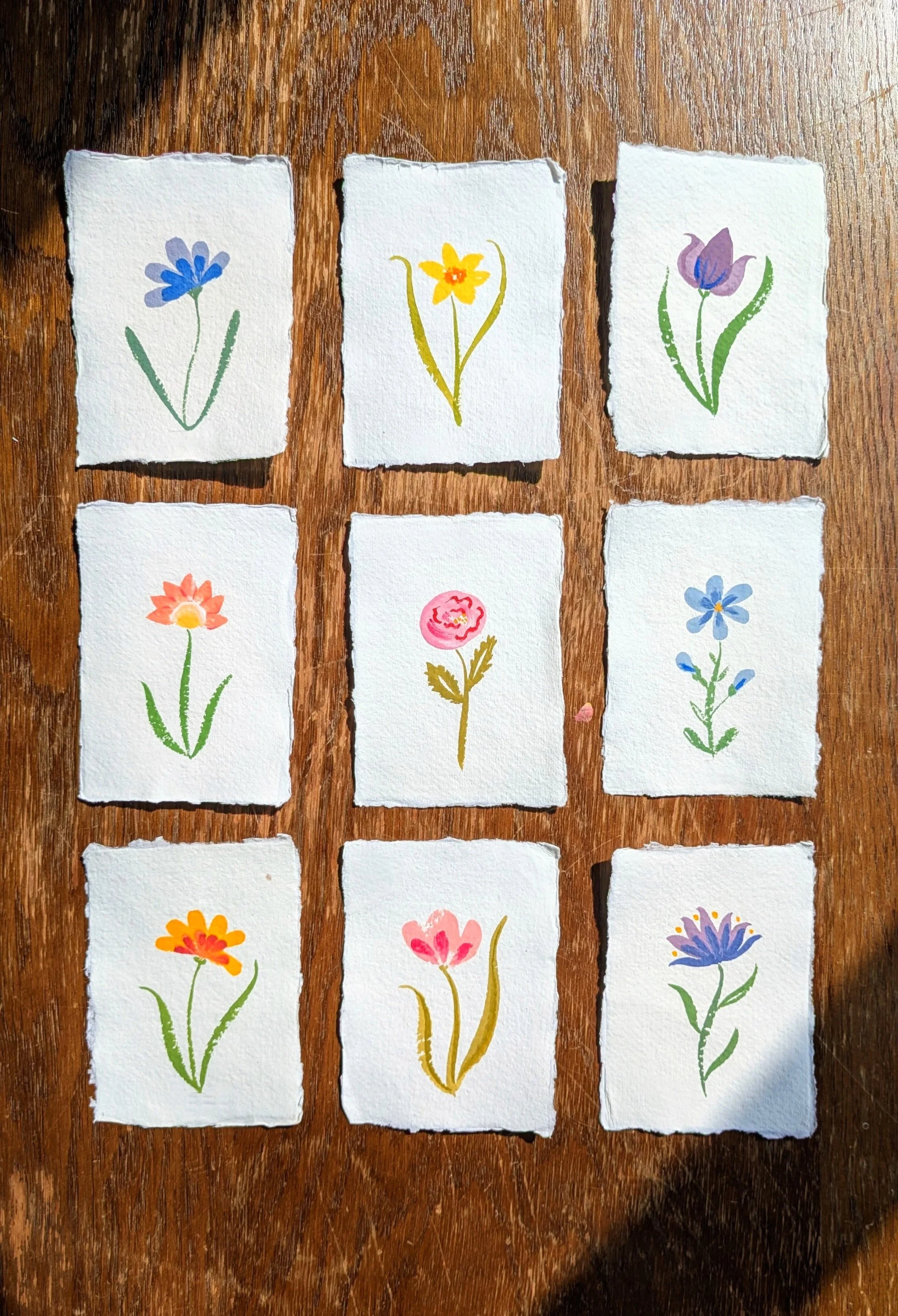 Watercolor paintings of various colorful flowers on torn white paper, laid out on a wooden surface.