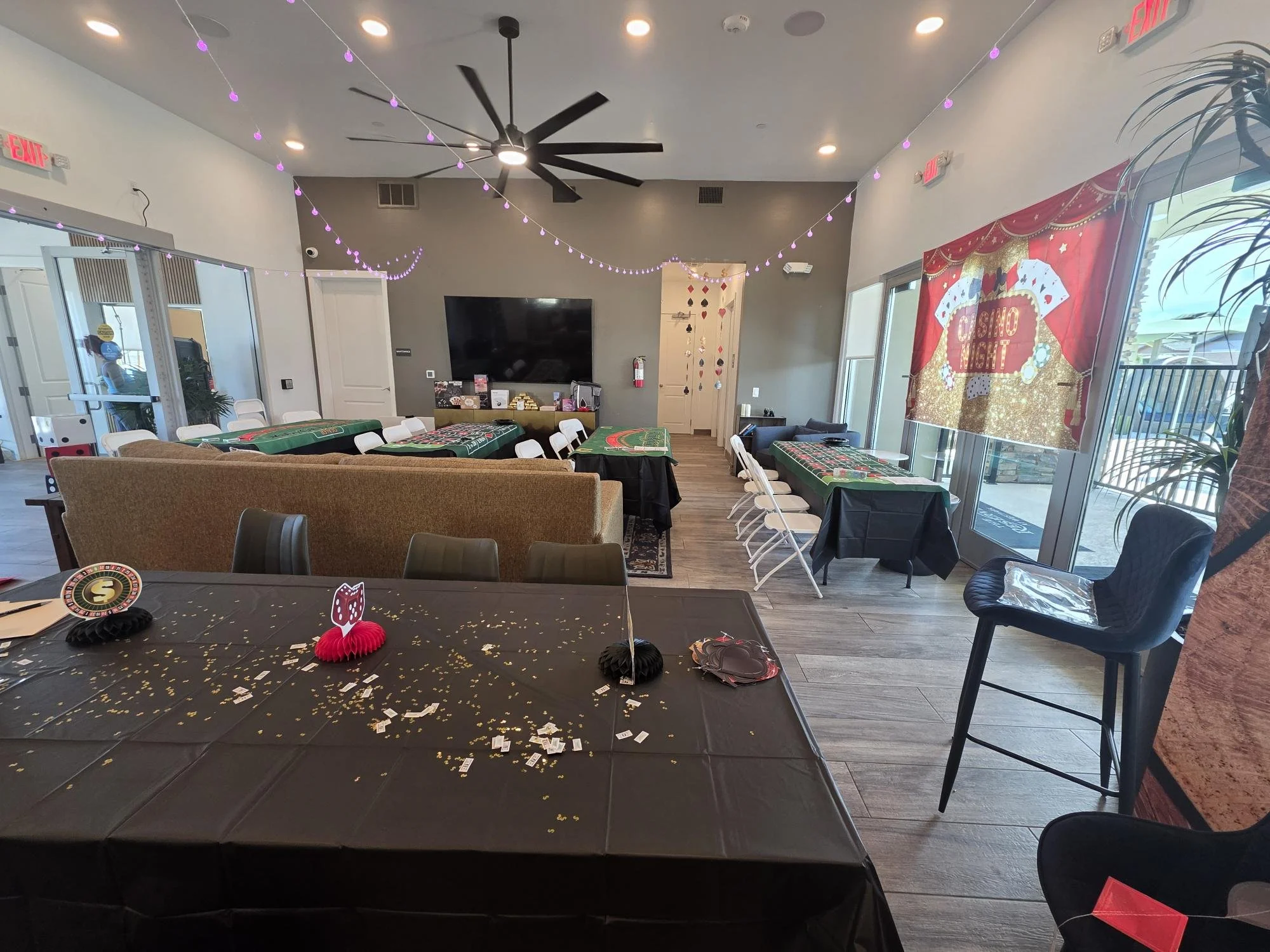 Interior of a casino-themed event room decorated with string lights, gaming tables covered with black cloths, and a large red and gold casino-themed banner on the wall that says 'Casino Night.' There are plastic chairs around the tables, a sofa, a large television, and a ceiling fan.