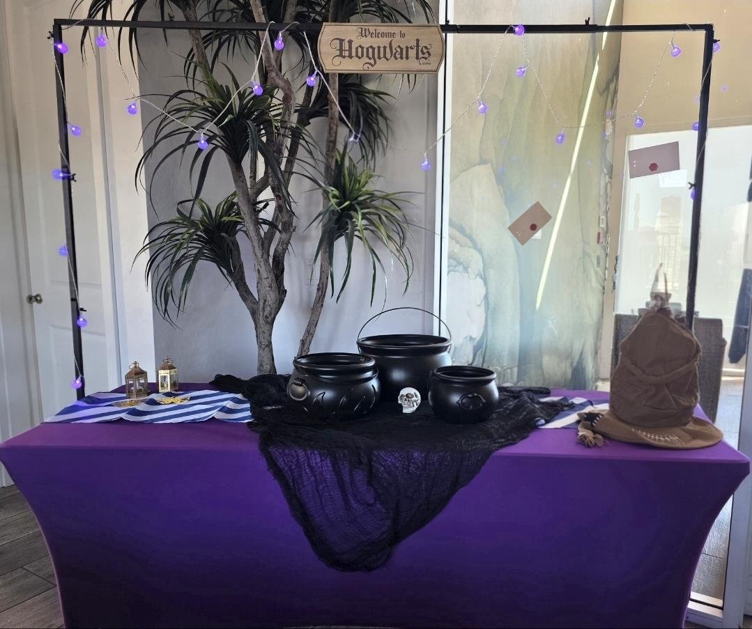 Hogwarts-themed display with purple tablecloth, black cauldrons, lanterns, a skull, and a sign that says "Welcome to Hogwarts," decorated with string lights and a large plant.