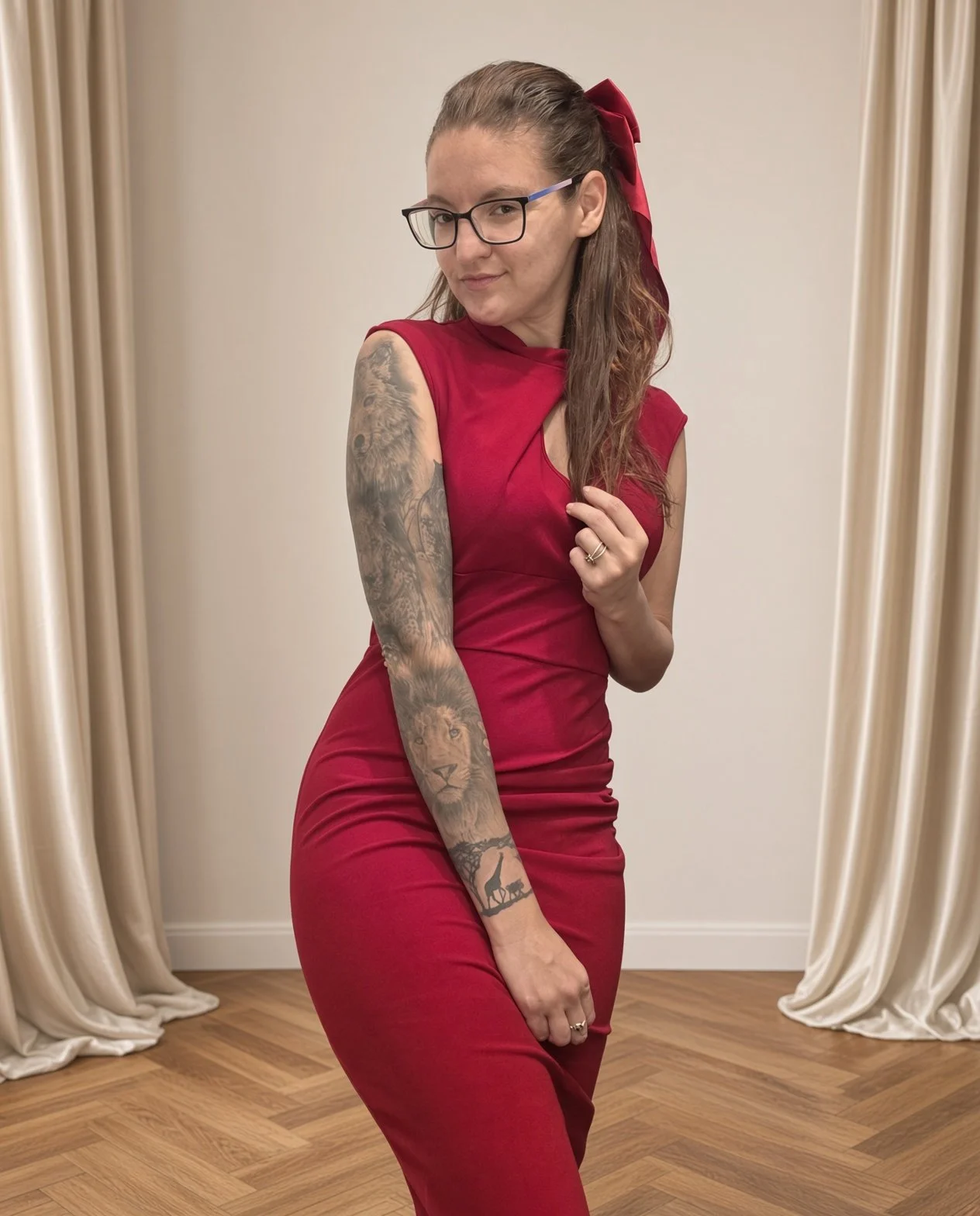 Woman wearing glasses and a red dress, showing her tattooed arm, standing in front of cream-colored curtains and wood flooring.