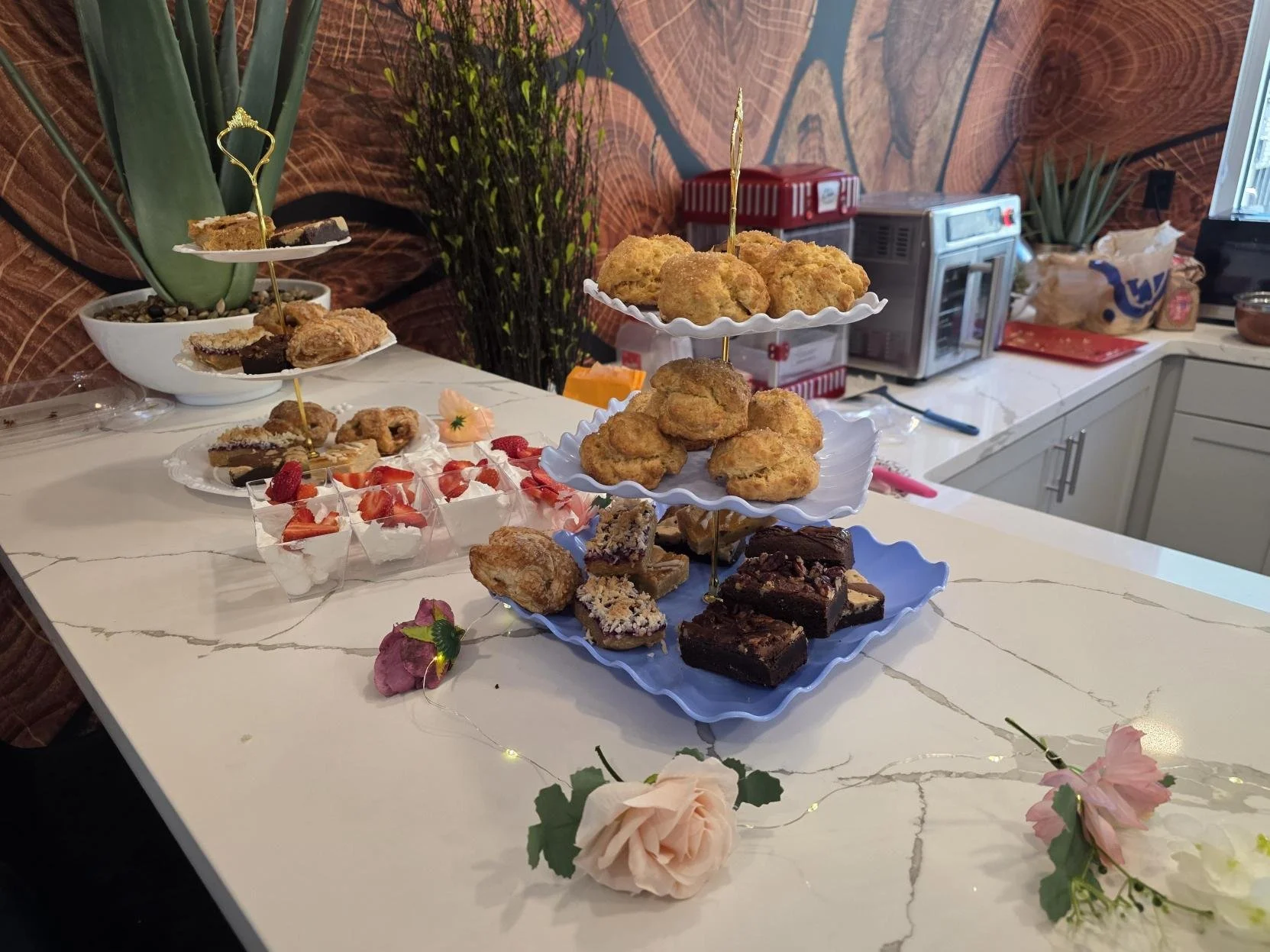 A table with assorted desserts including cookies, brownies, and strawberries, decorated with flowers and fairy lights, in a kitchen with wrapped gifts and plants in the background.