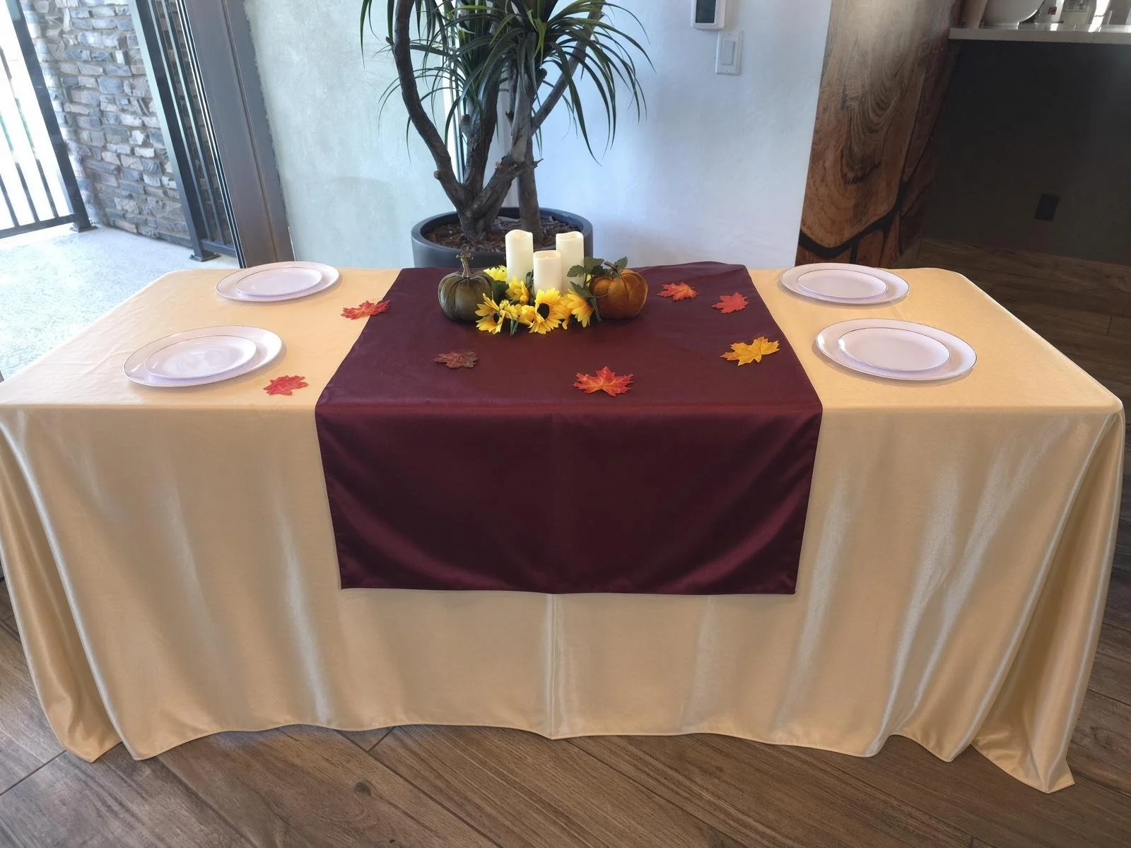 A table with a cream-colored tablecloth, decorated with a maroon table runner, an autumn centerpiece of small pumpkins, sunflowers, and candles, and scattered fall leaves. The table is set for four with white plates, and there is a large potted plant next to the table with a sliding glass door and outdoor space visible in the background.