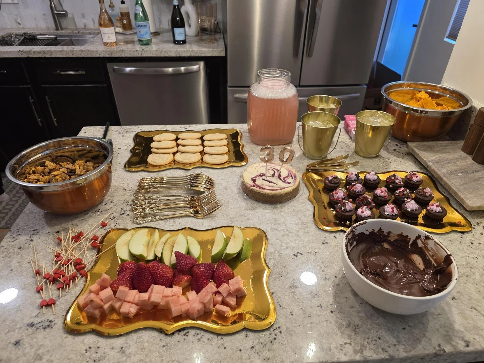 A kitchen countertop with various snacks and desserts for a celebration, including cupcakes, fruit, a small cake, chocolate, candies, and drinks.