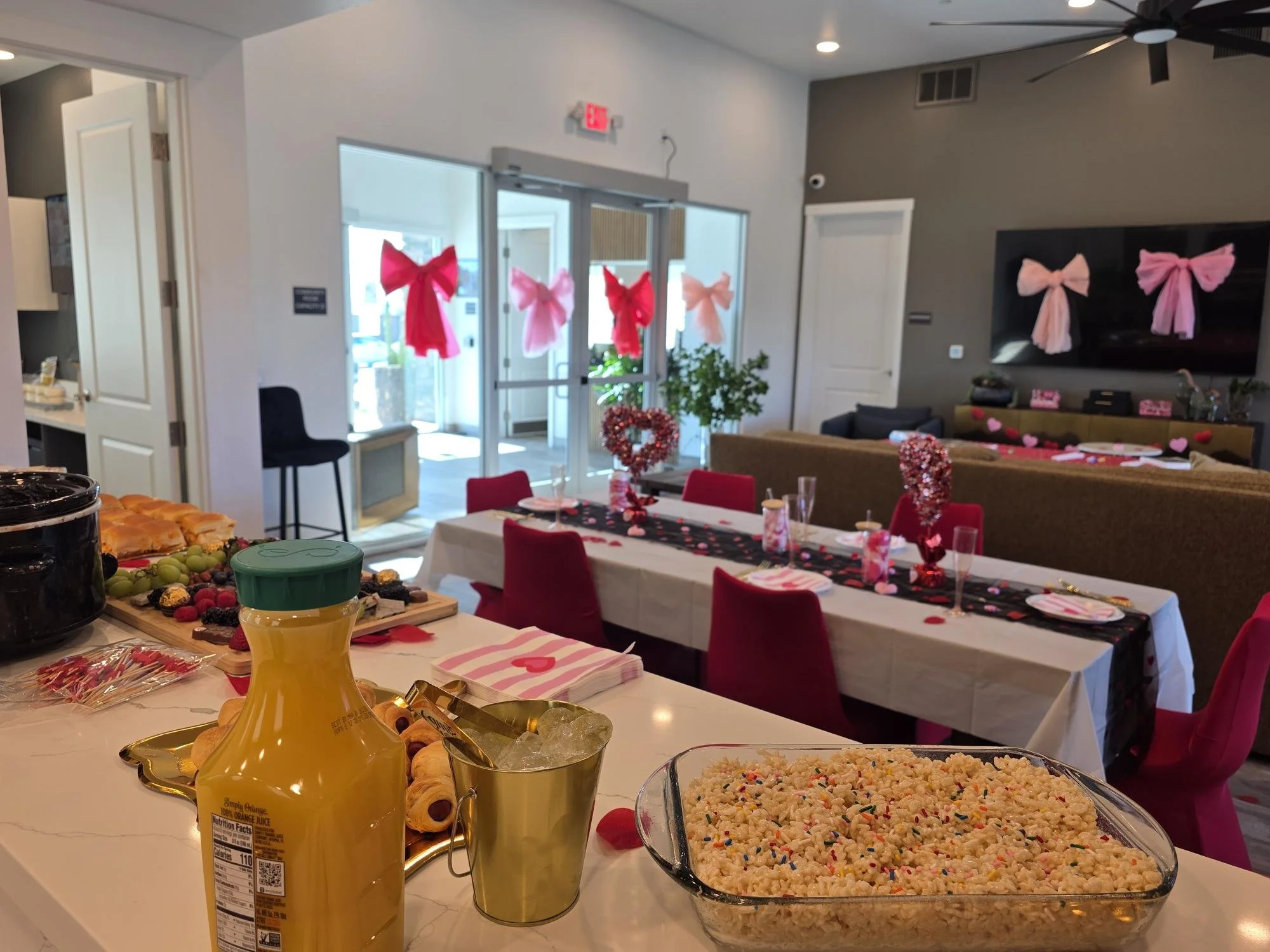 Decorative Valentine's Day themed party with pink bows on the wall and table settings, featuring snacks and drinks, in a modern room with computer monitor and sliding glass door.