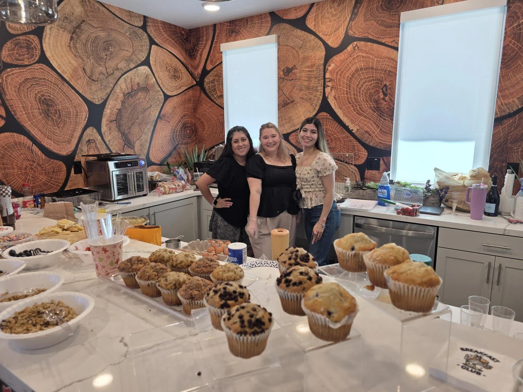 Three women standing in a kitchen with a table full of cupcakes and baked goods, smiling at the camera. The kitchen has a wood slice pattern wallpaper, white cabinets, and various baking supplies on the counter.