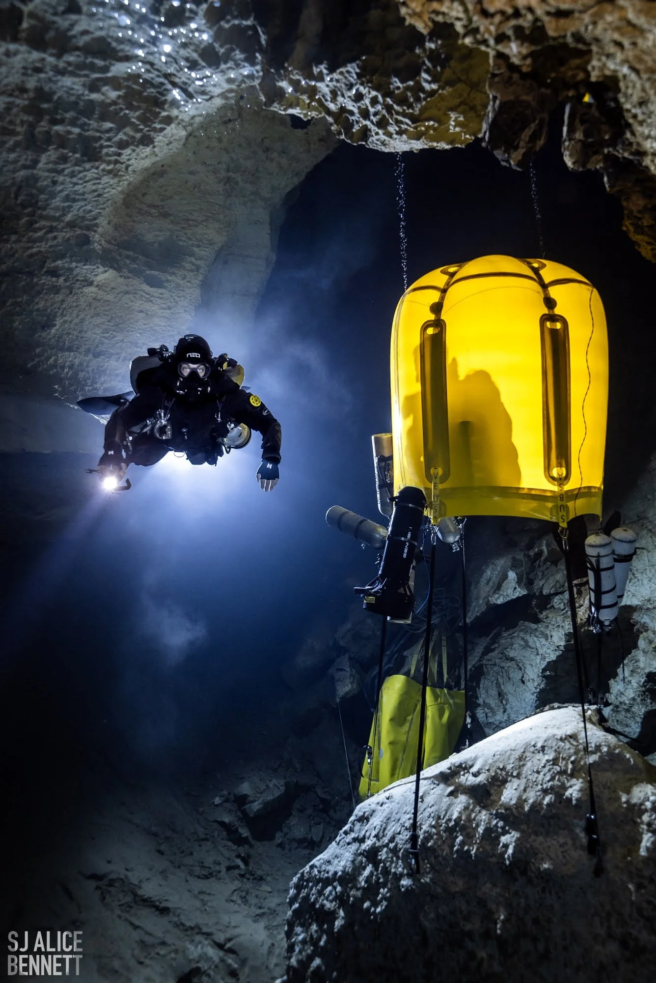 A person in scuba diving gear inside a dark cave, illuminated by a flashlight, with a yellow underwater exploration equipment nearby and stalactites hanging from the ceiling.