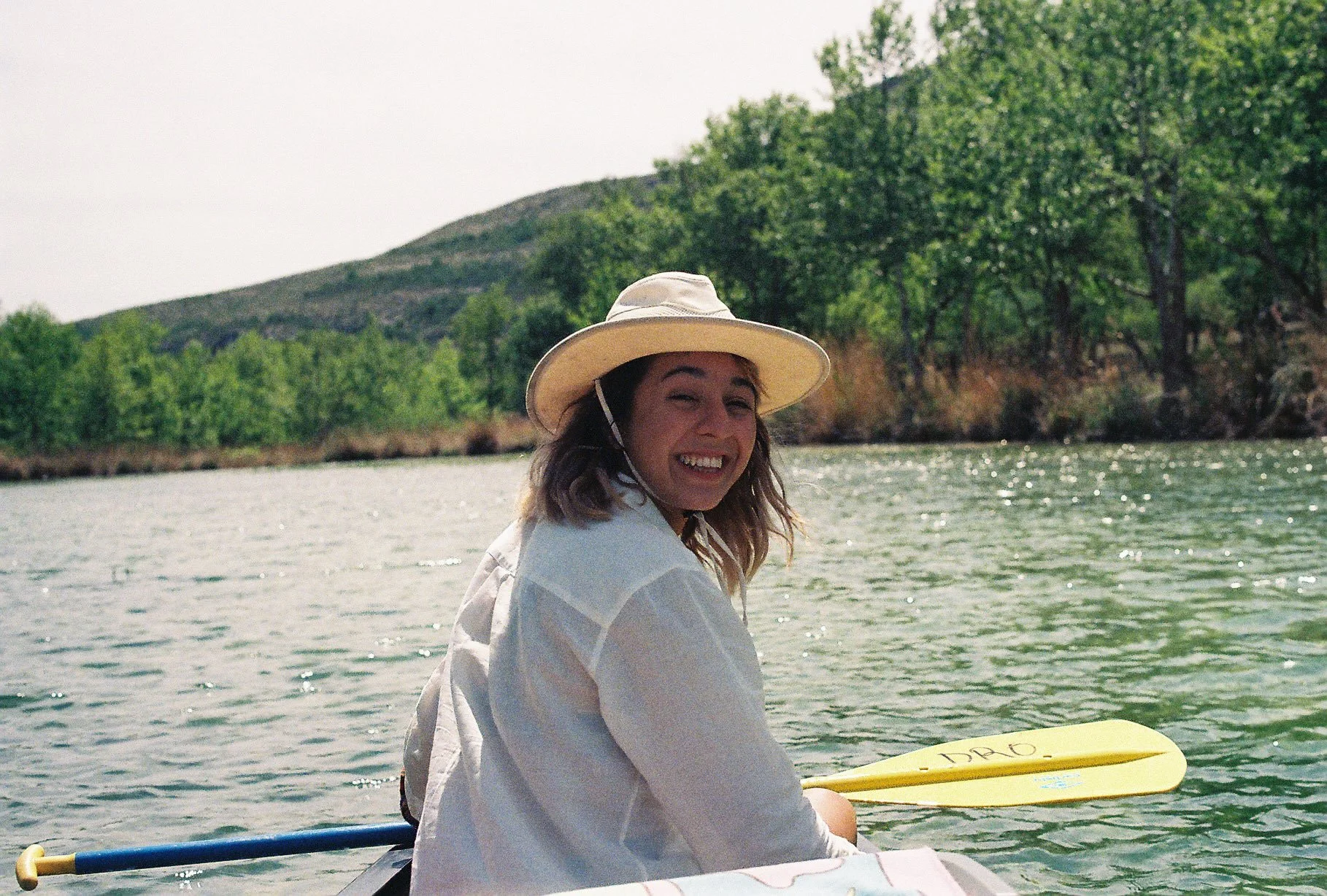 A woman sitting in a boat on a river, smiling, wearing a wide-brimmed hat and a white jacket, holding a yellow paddle, with green trees and a hill visible in the background.