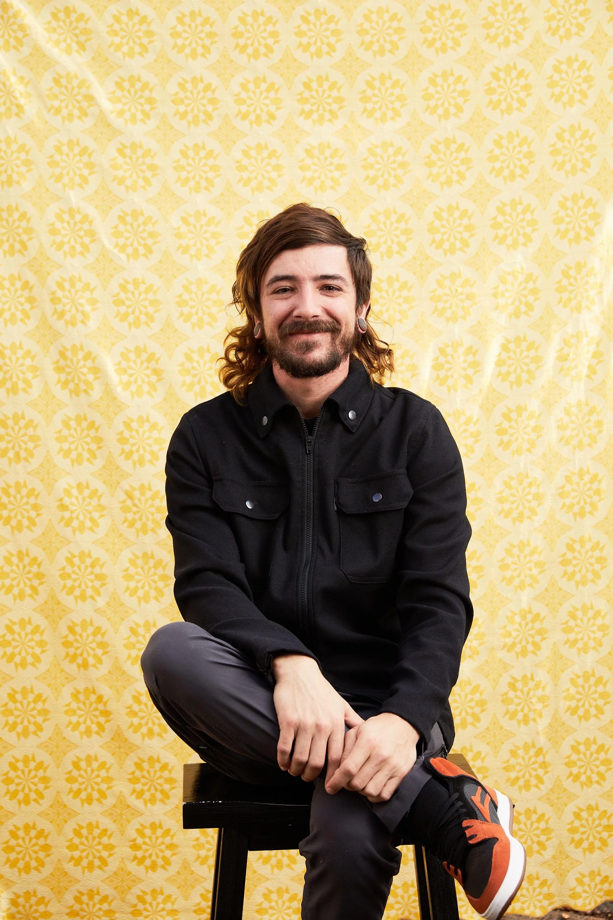 Portrait of a man with long brown hair, beard, and hearing pipes, sitting on a black stool in front of a yellow patterned background, wearing a black jacket, gray pants, and black and orange sneakers.