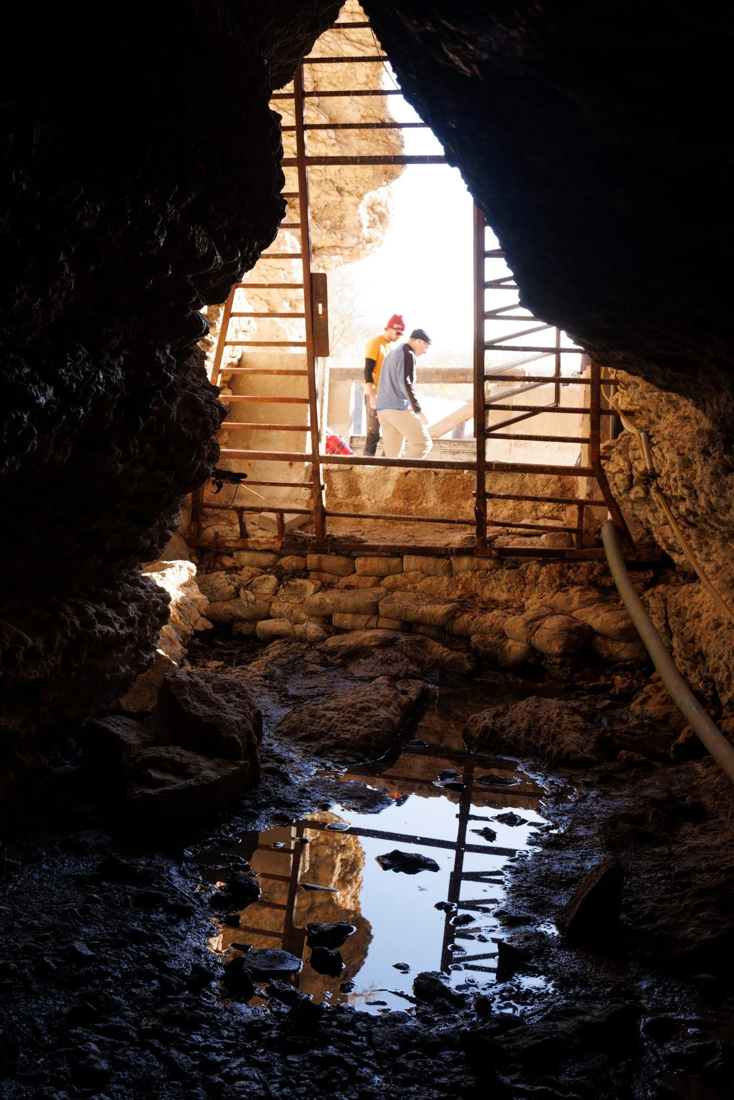 View from inside a narrow cave or tunnel looking toward a sunny outdoor scene with two people, one in a yellow shirt and one in a gray jacket, standing near a wooden railing and ladder. A small body of water and rocks are visible at the cave's entrance.