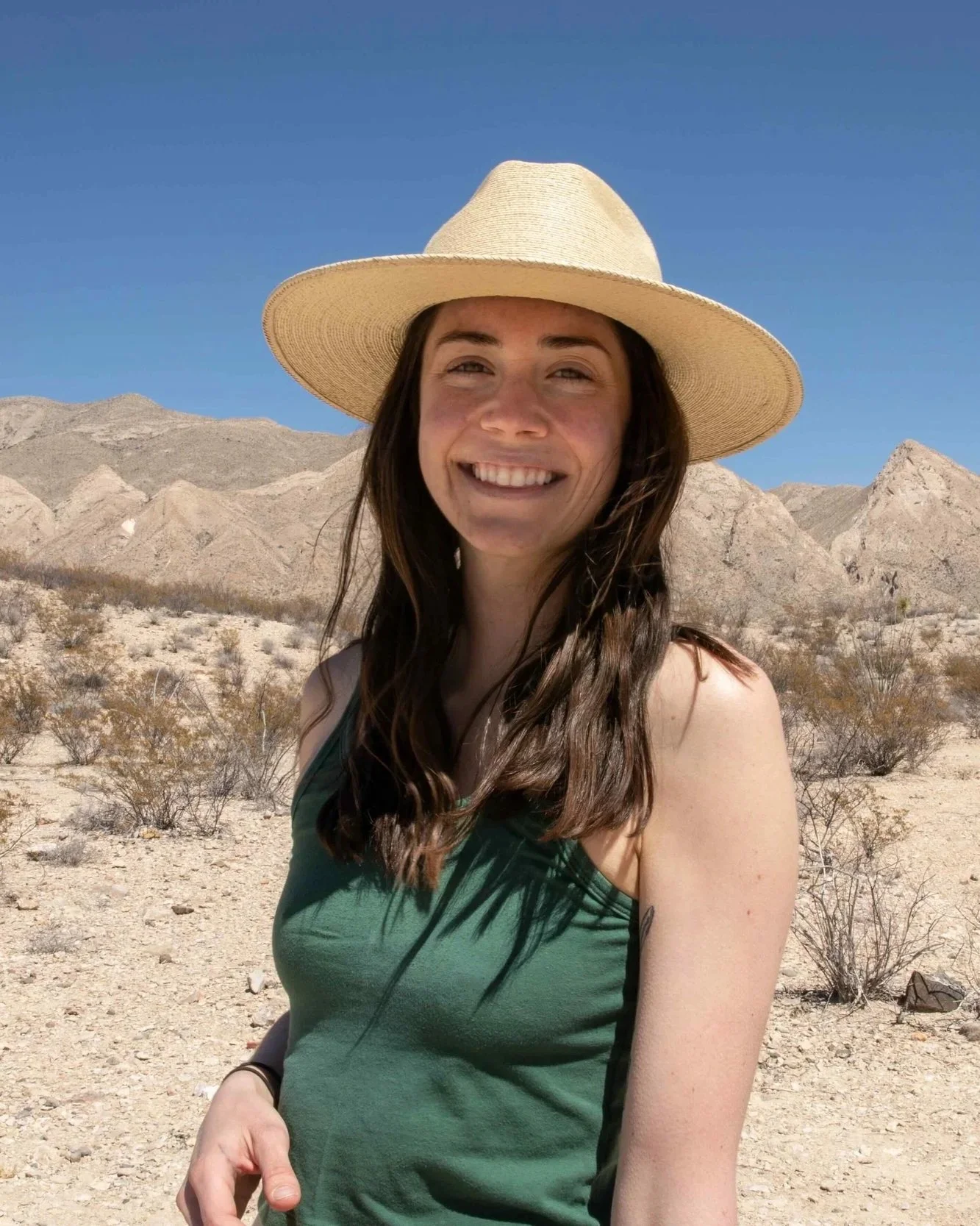 A woman with long brown hair wearing a large sunhat and green tank top, smiling in a desert landscape with mountains and sparse vegetation, under a clear blue sky.