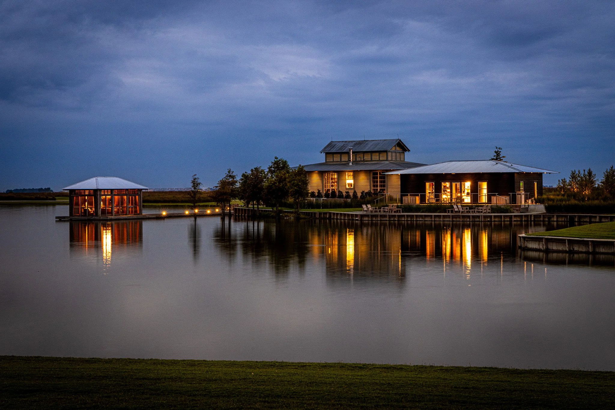 A house with large windows and a deck illuminated from within, situated beside a calm body of water. The house is surrounded by greenery, and smaller structures are visible nearby. The sky is cloudy and dark, indicating evening or night.