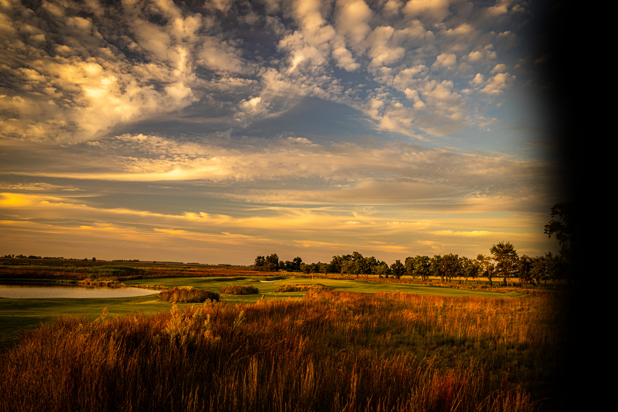 A scenic view of a landscape during sunset with a partly cloudy sky, grassy fields, and trees.