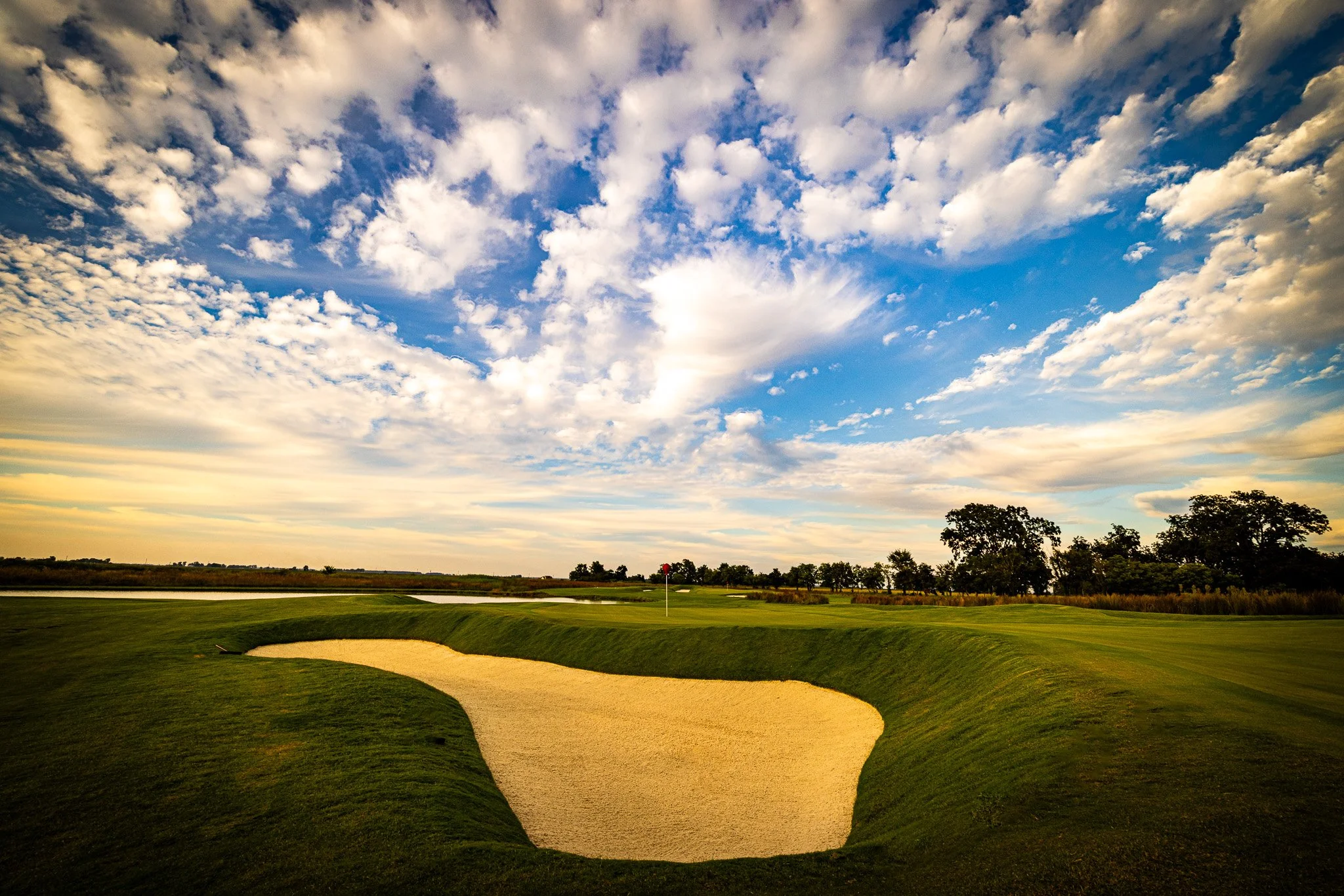 A golf course with a sand trap and green grass under a partly cloudy sky with trees in the distance.