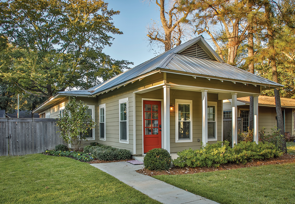 A single-story house with beige siding and a metal roof, featuring a front porch with white columns, multiple windows with white frames, and a red front door. The yard has neatly trimmed grass, bushes, and a small garden, with a concrete walkway lead
