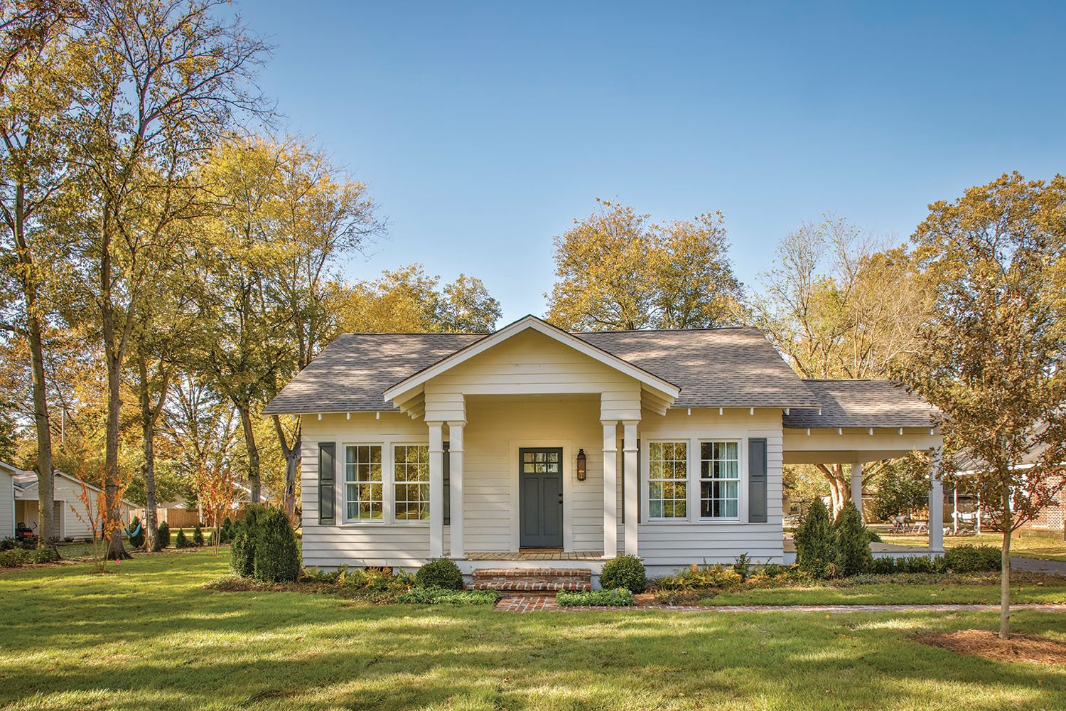 Front view of a white house with gray shutters and a small front porch with columns, surrounded by green lawn and trees, under a clear blue sky.