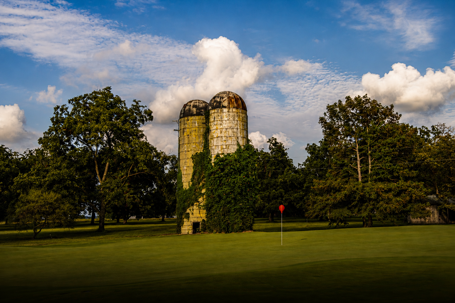 Two old silos surrounded by trees on a grassy area under a partly cloudy sky, with a golf flag in the foreground.