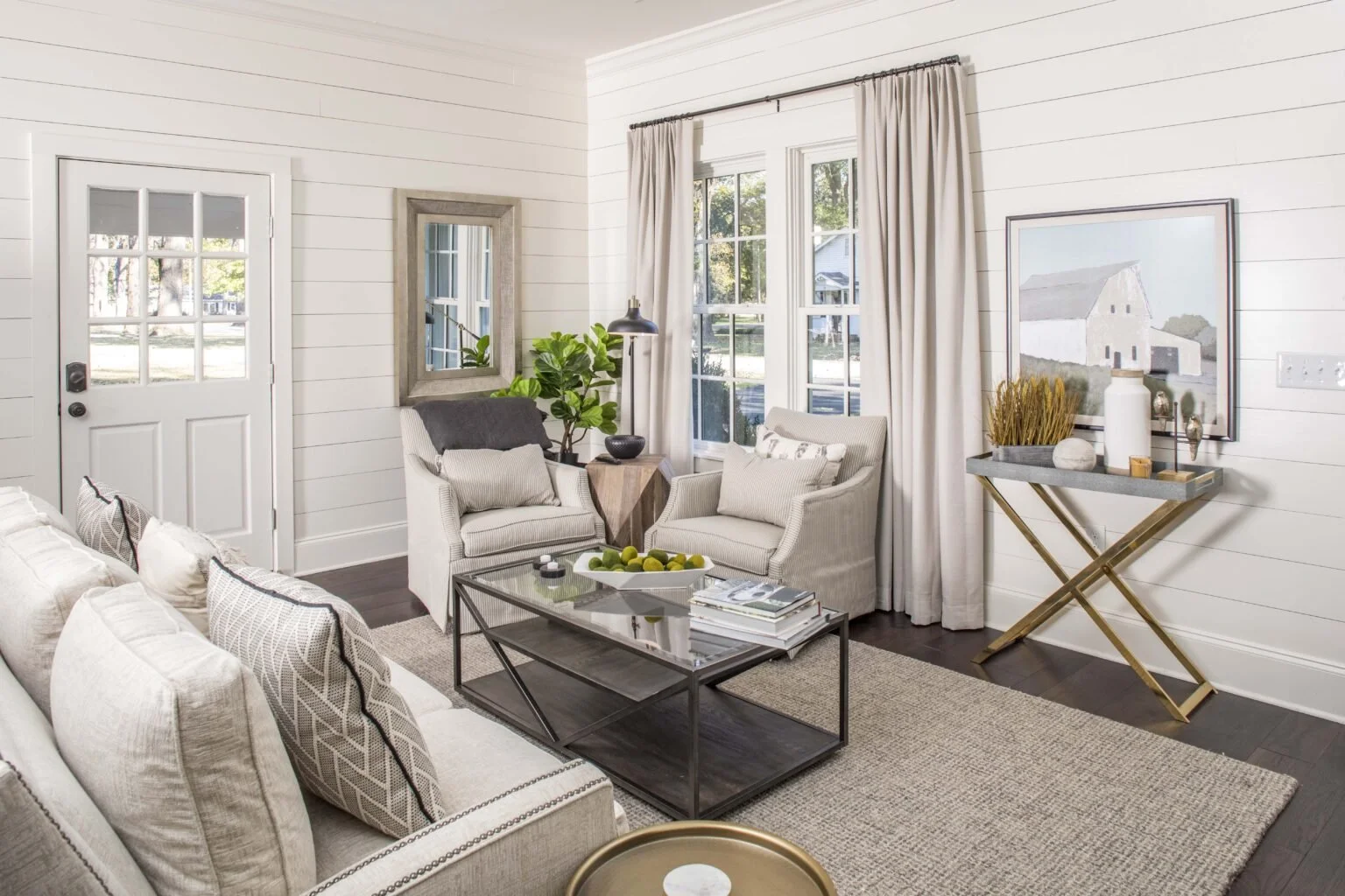 Bright living room with white shiplap walls, beige curtains, two white armchairs, a sofa with patterned cushions, a glass coffee table with a tray of green apples, and a small side table with a potted plant; a side console with decor items and a larg