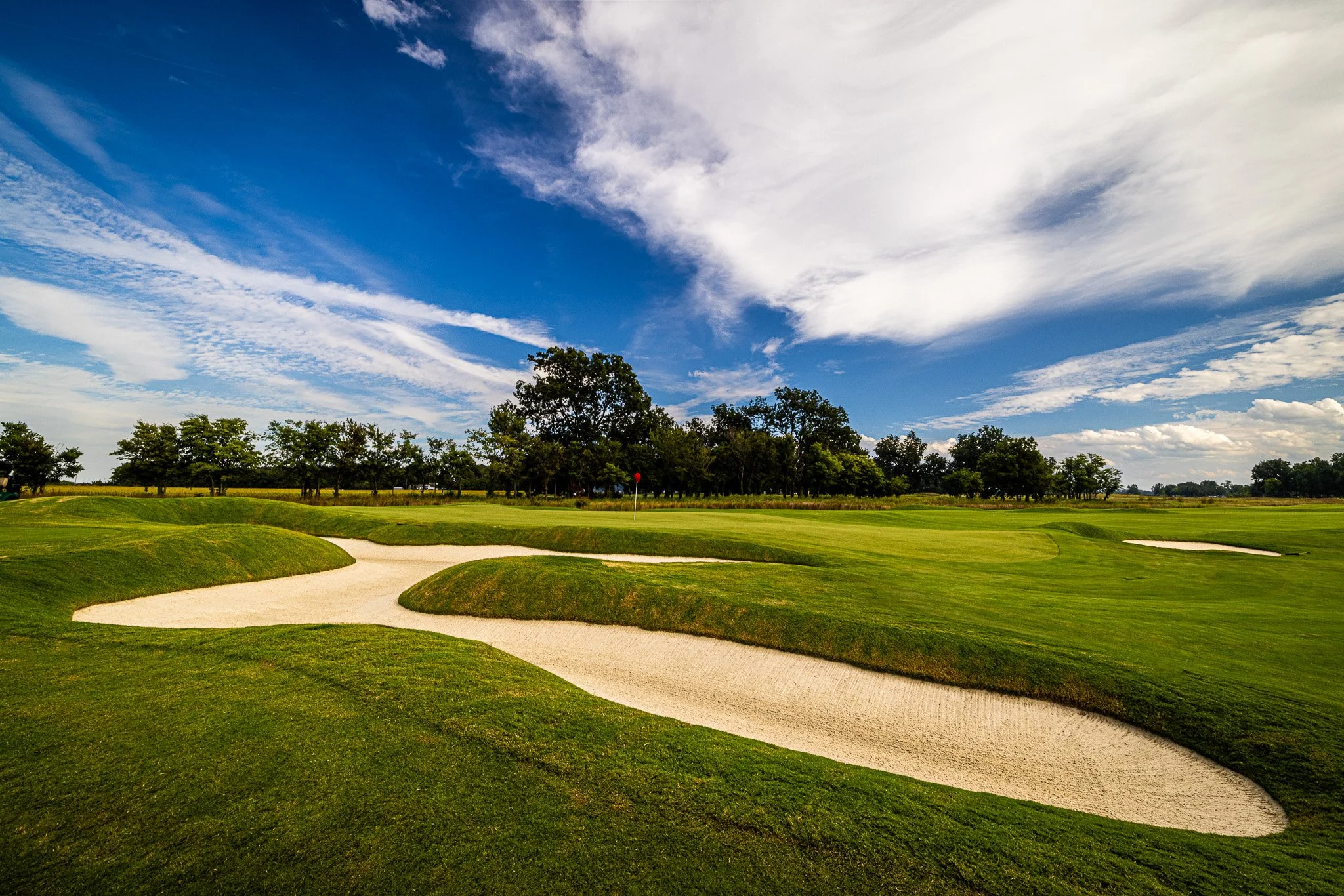 A golf course with sand traps, a putting green, and a flagstick, surrounded by green grass and trees under a partly cloudy sky.
