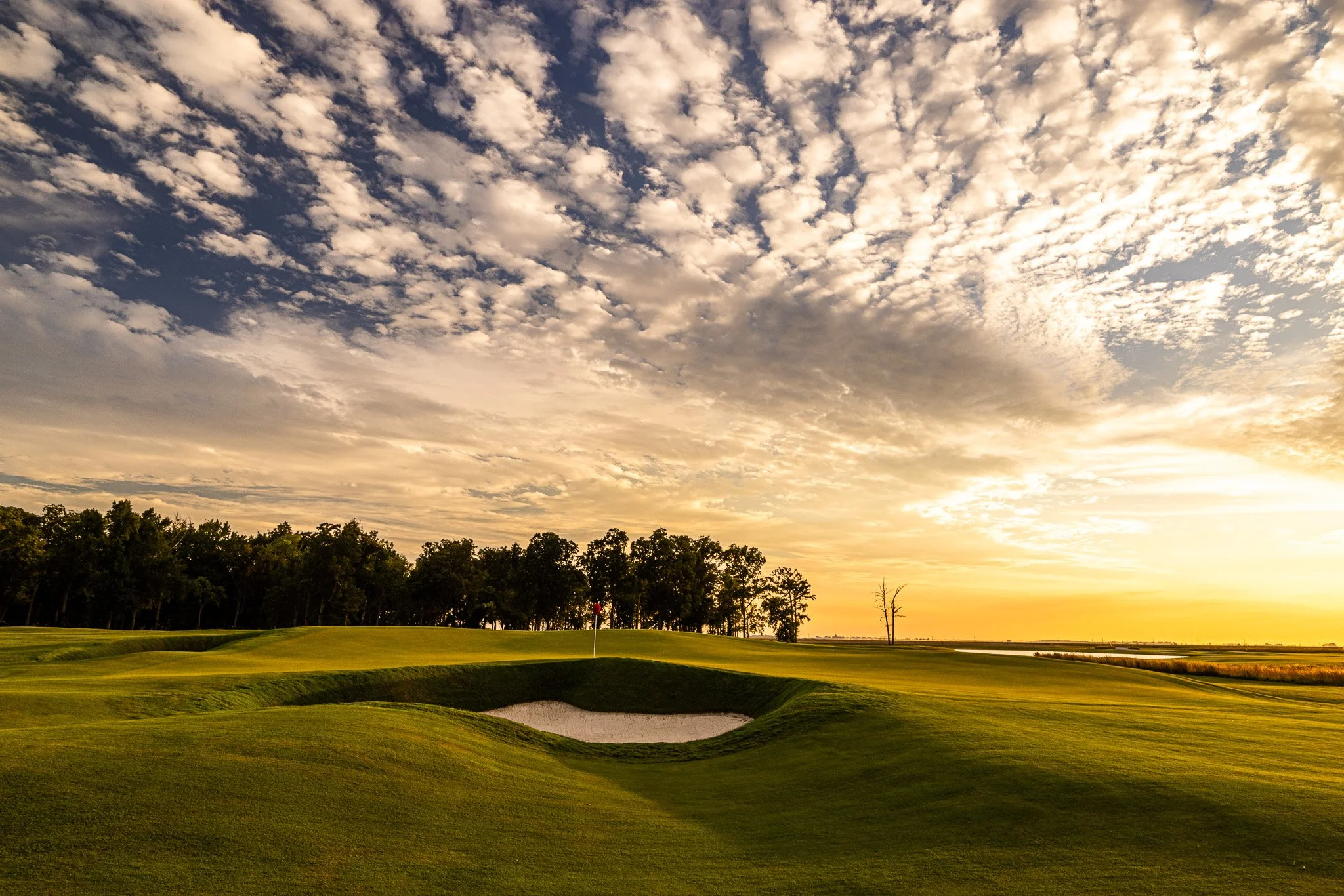 A golf course at sunset with green grass, sand trap, trees, and a flag on the green, under a sky filled with scattered clouds and a warm golden glow.
