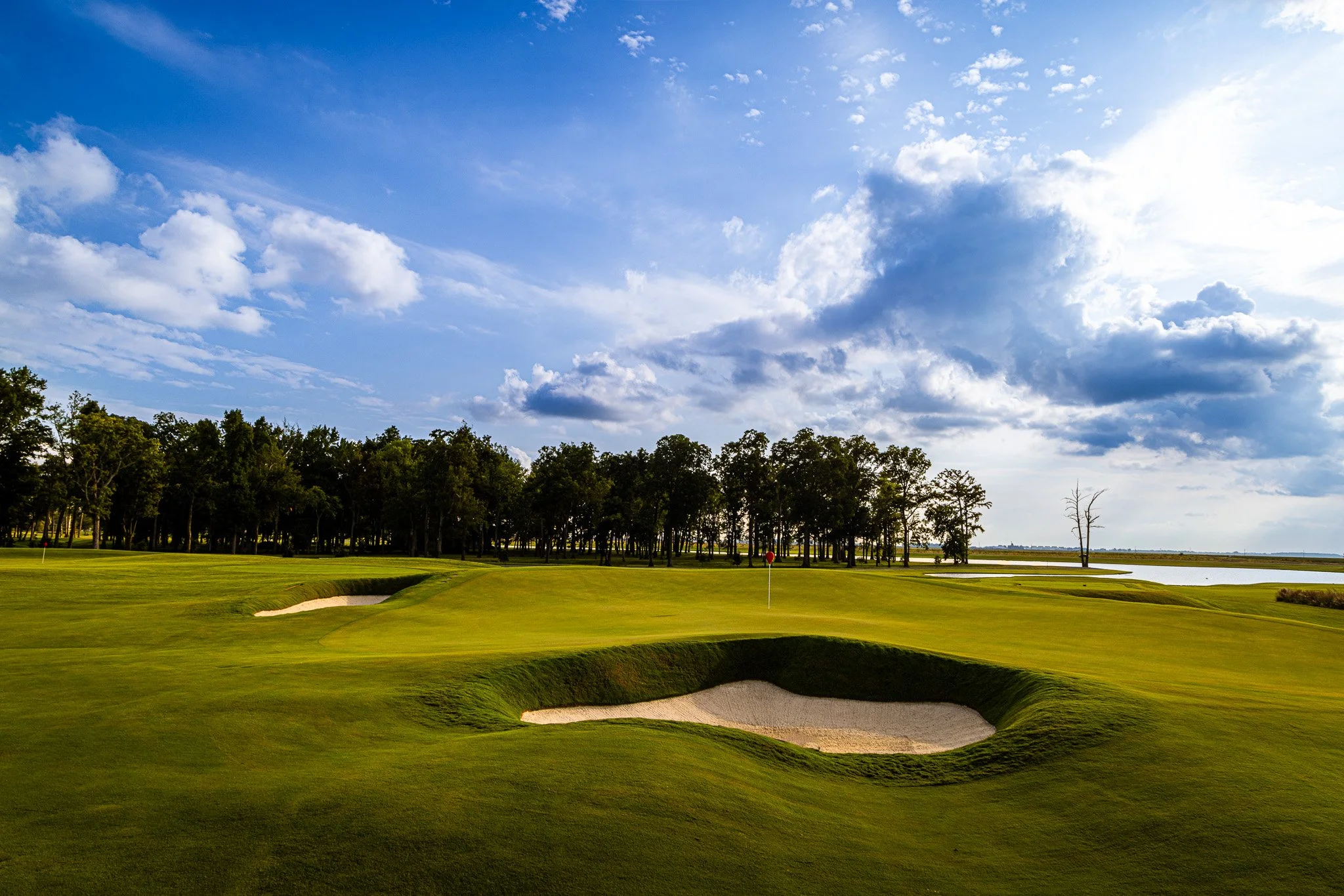 A golf course with green grass, sand bunkers, a putting green with a flag, and a background of trees under a partly cloudy sky.