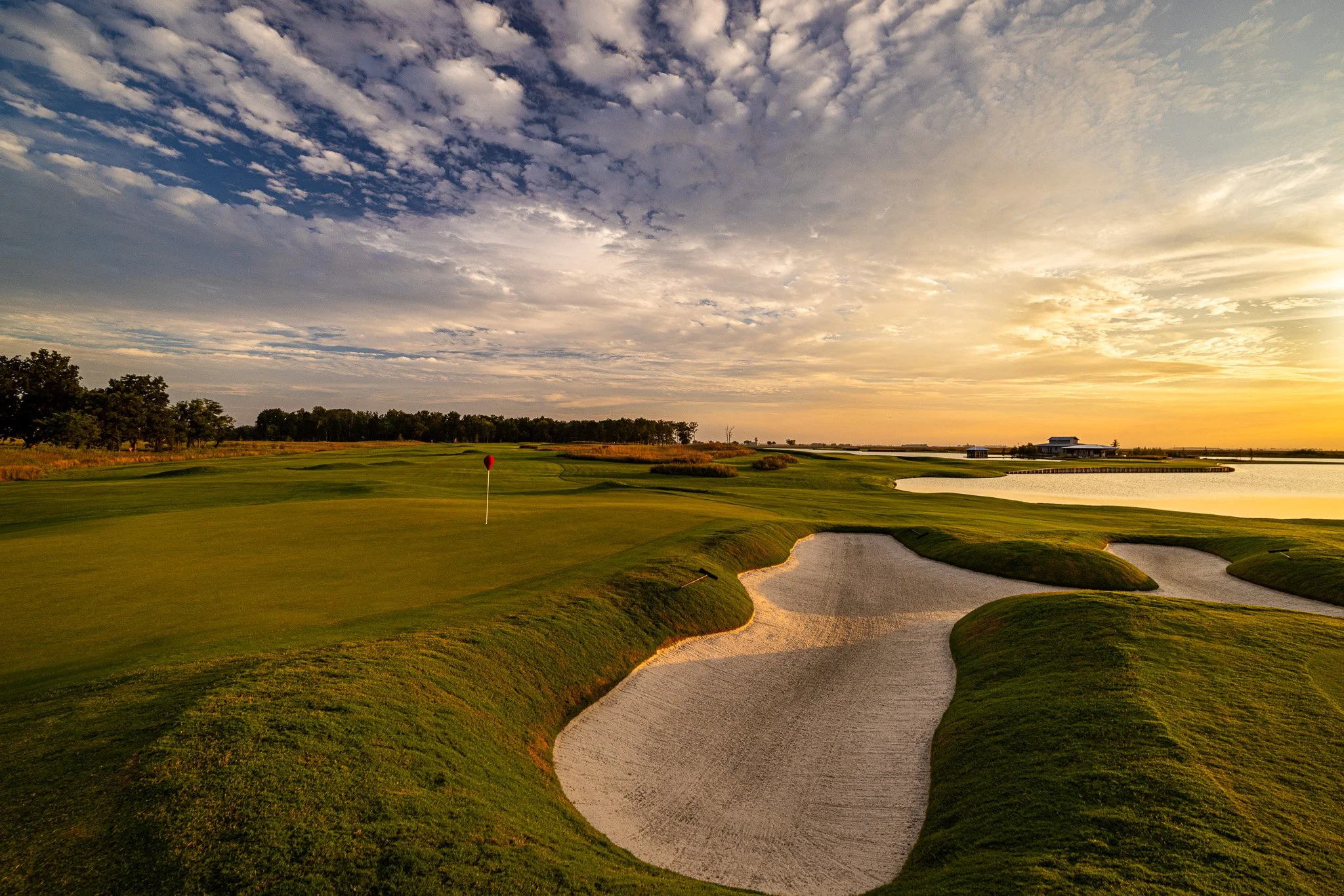 A golf course during sunset with green grass, sand traps, a body of water, and a sky filled with clouds.