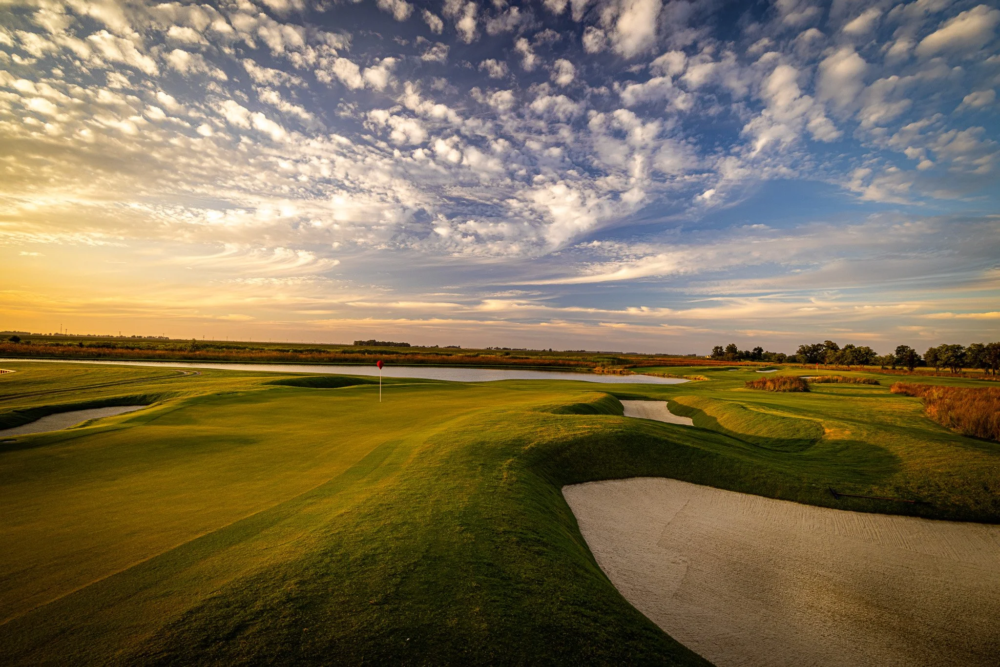 A scenic view of a golf course during sunset with a clear sky filled with clouds, featuring a pond, sand traps, and green fairways.