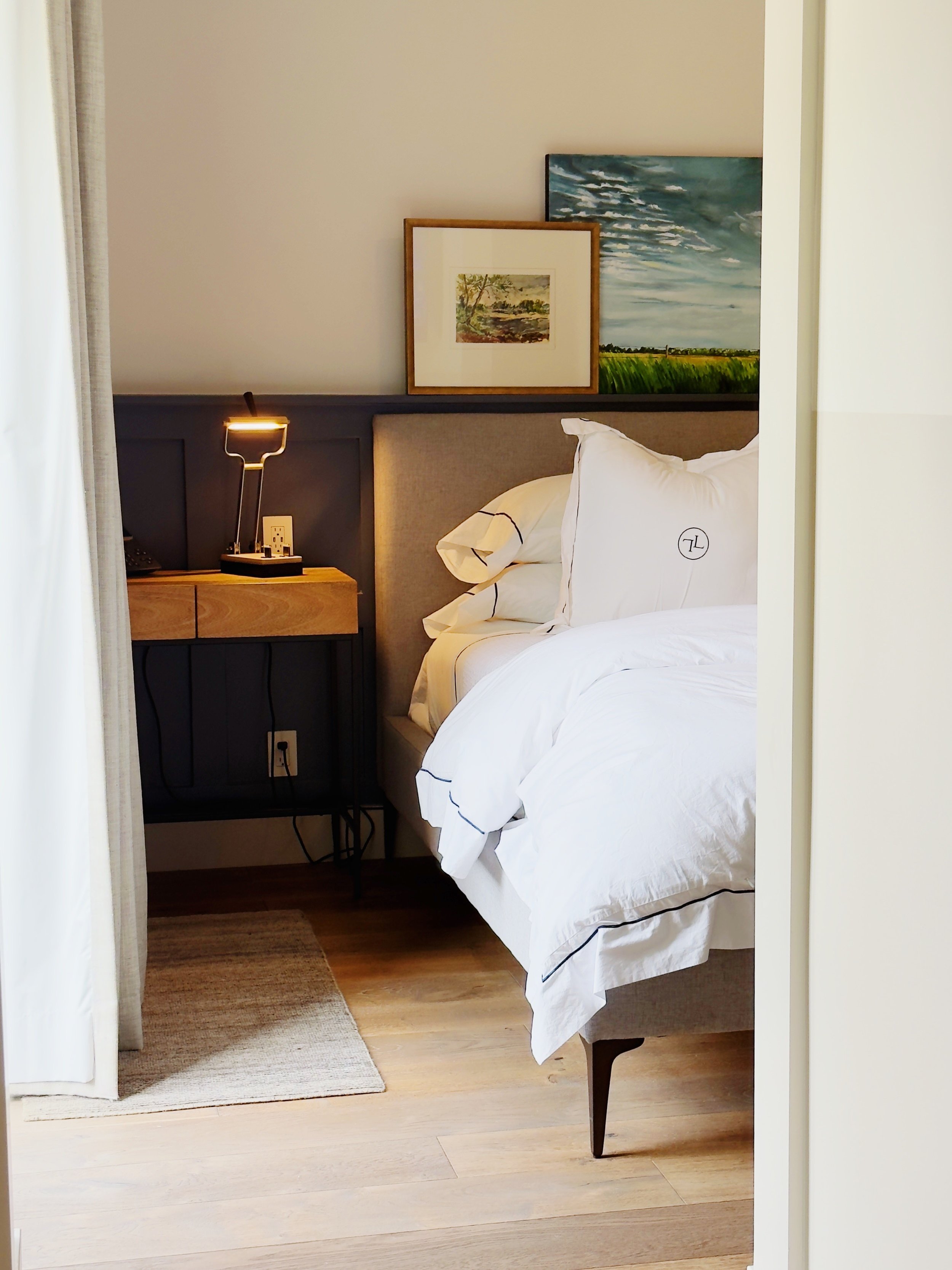 A hotel bedroom with a white bed, bedside table, lamp, framed artwork, and wooden flooring.