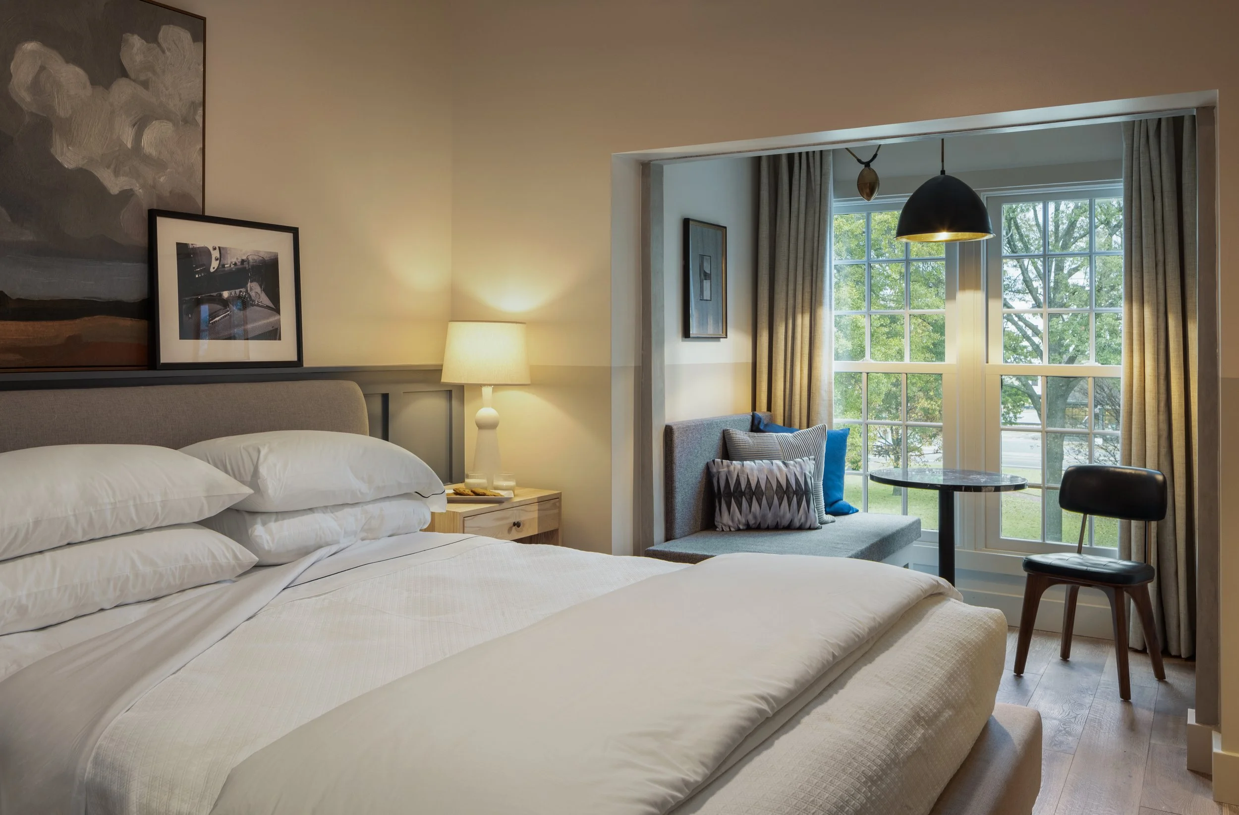 A cozy bedroom with a bed, side table, lamp, framed artwork, and window with curtains and outdoor greenery visible.