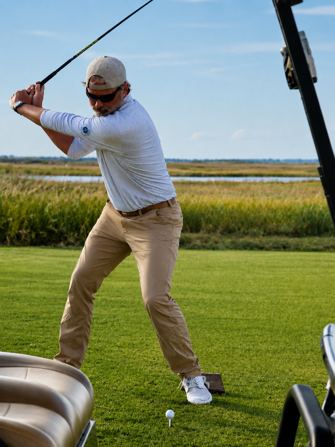 Man with a beard and sunglasses in a baseball cap taking a golf swing on a golf course with green grass, water and fields in the background.