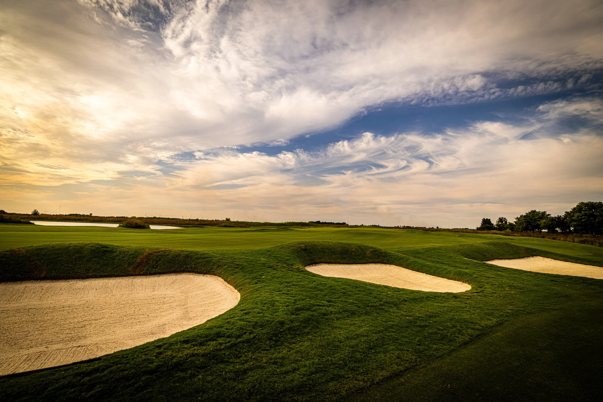 A golf course with sand bunkers, green grass, and a backdrop of trees and a partly cloudy sky.