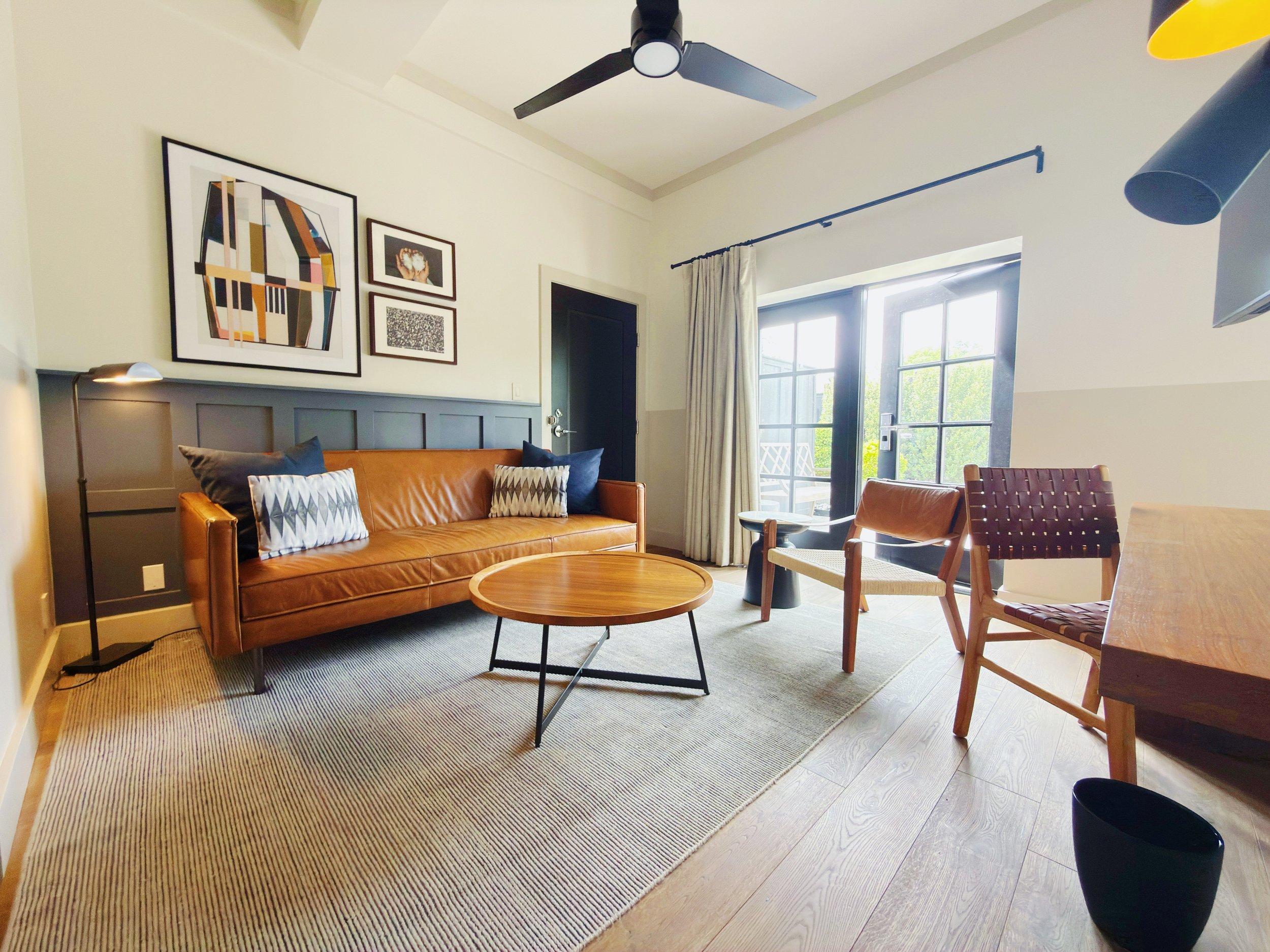 A living room with an orange leather sofa, a round wooden coffee table, a beige and black striped rug, multiple framed pictures on the wall, a black ceiling fan, large windows with white curtains, and a pair of wooden chairs with woven seats.