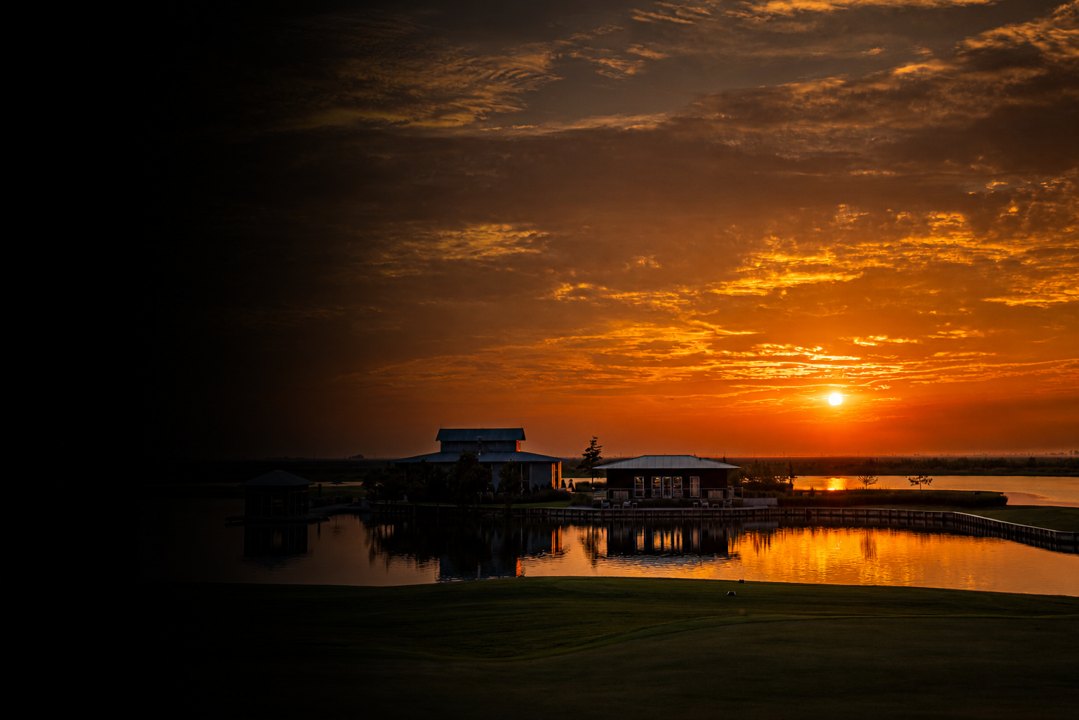 A sunset over a farm with buildings, trees, and a body of water reflecting the sky.