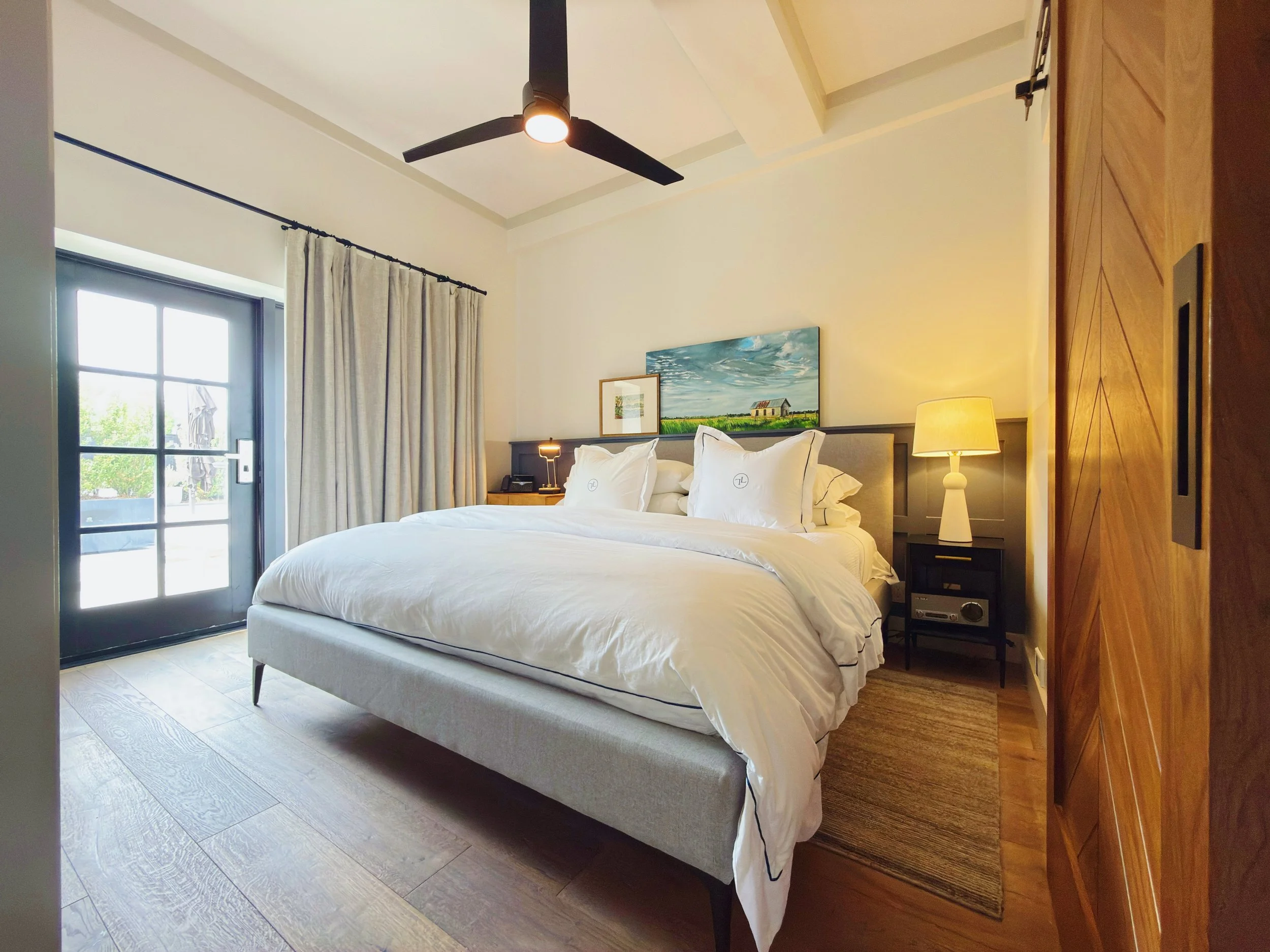 A neatly made bed with white linens and pillows in a modern bedroom with a window and sliding glass door, a nightstand with a lamp, and artwork above the bed.
