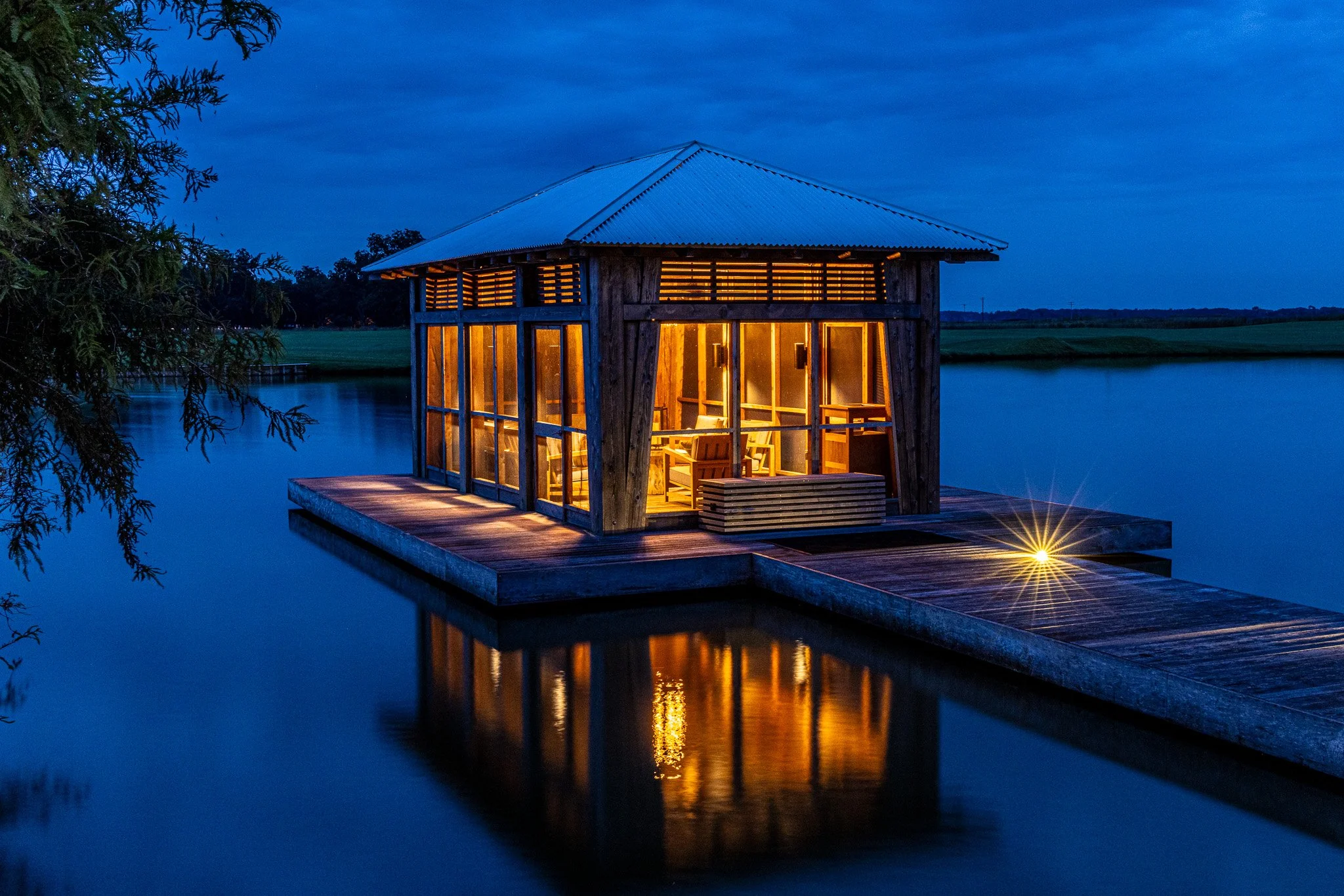 A wooden lake house with large glass windows and a corrugated metal roof, illuminated warmly from the inside, situated on a dock over a calm lake during dusk with blue sky and trees in the background.