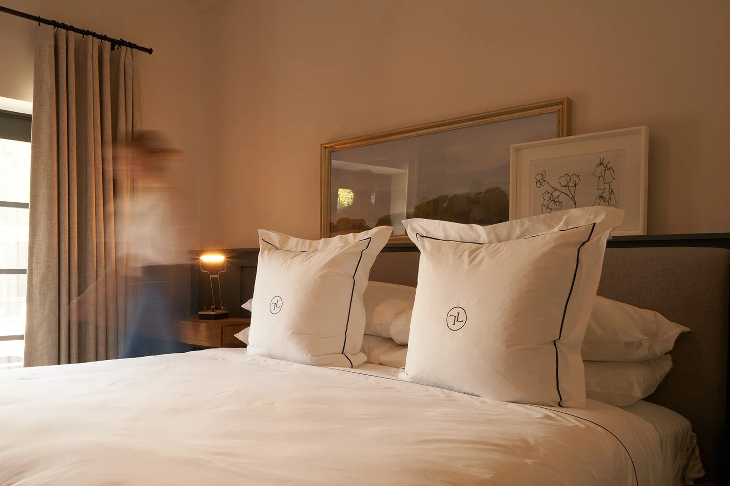 A bedroom with a bed, two large pillows with a logo, framed artwork on the wall, a window with beige curtains, and a person walking past, appearing blurred.