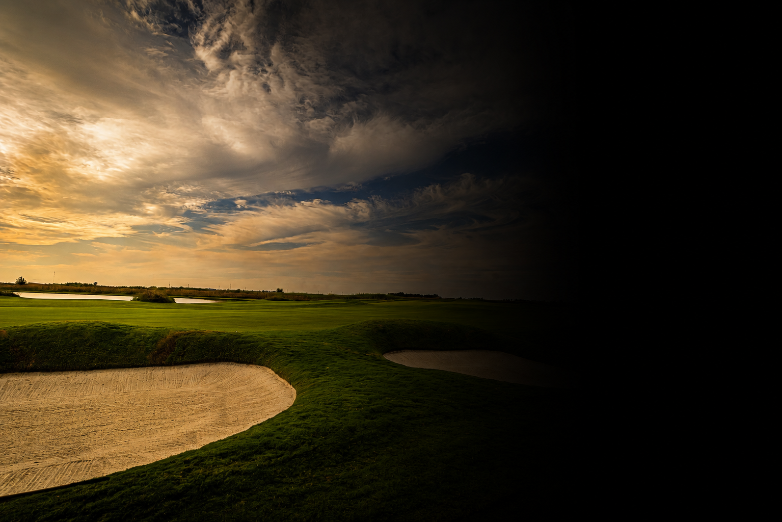 A landscape with partly cloudy skies, green grass, and sand bunkers on a golf course during late afternoon or early evening.