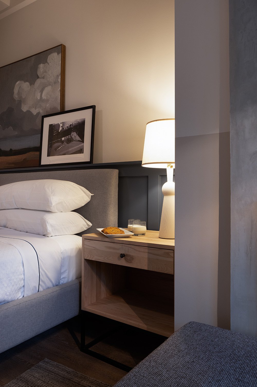 A cozy bedroom with a bed, pillows, framed artwork on the wall, a wooden nightstand with a lamp, a plate of cookies, and a glass of milk.