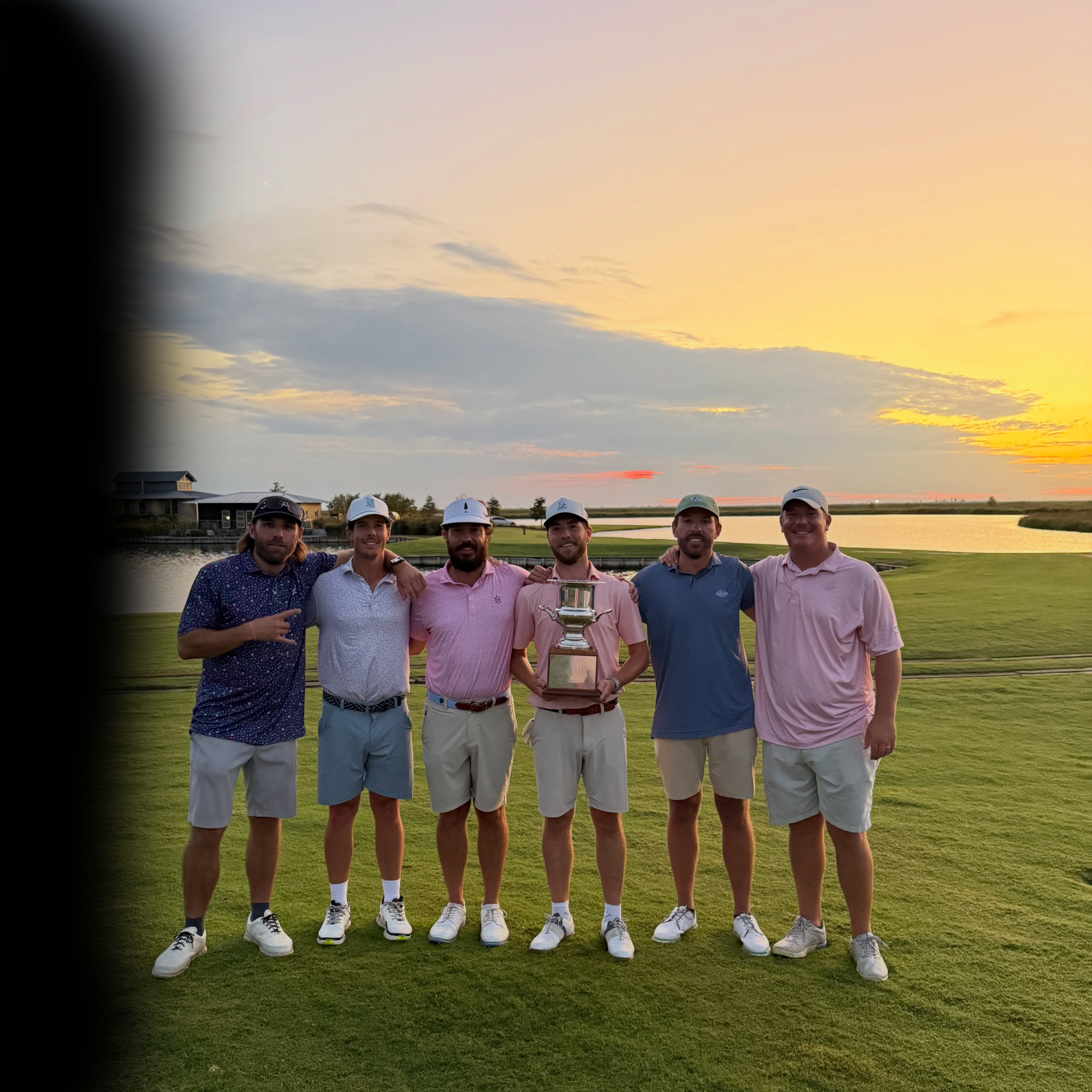 A group of seven men in golf attire standing together on a golf course at sunset, holding a trophy. The sky is partly cloudy with a pink and orange hue.