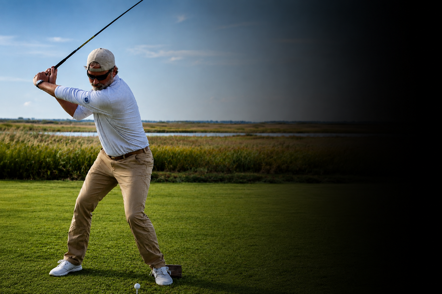An older man wearing a white cap, sunglasses, a white long-sleeve shirt, and khaki pants is playing golf on a lush green course near a marshy area, about to swing a golf club.