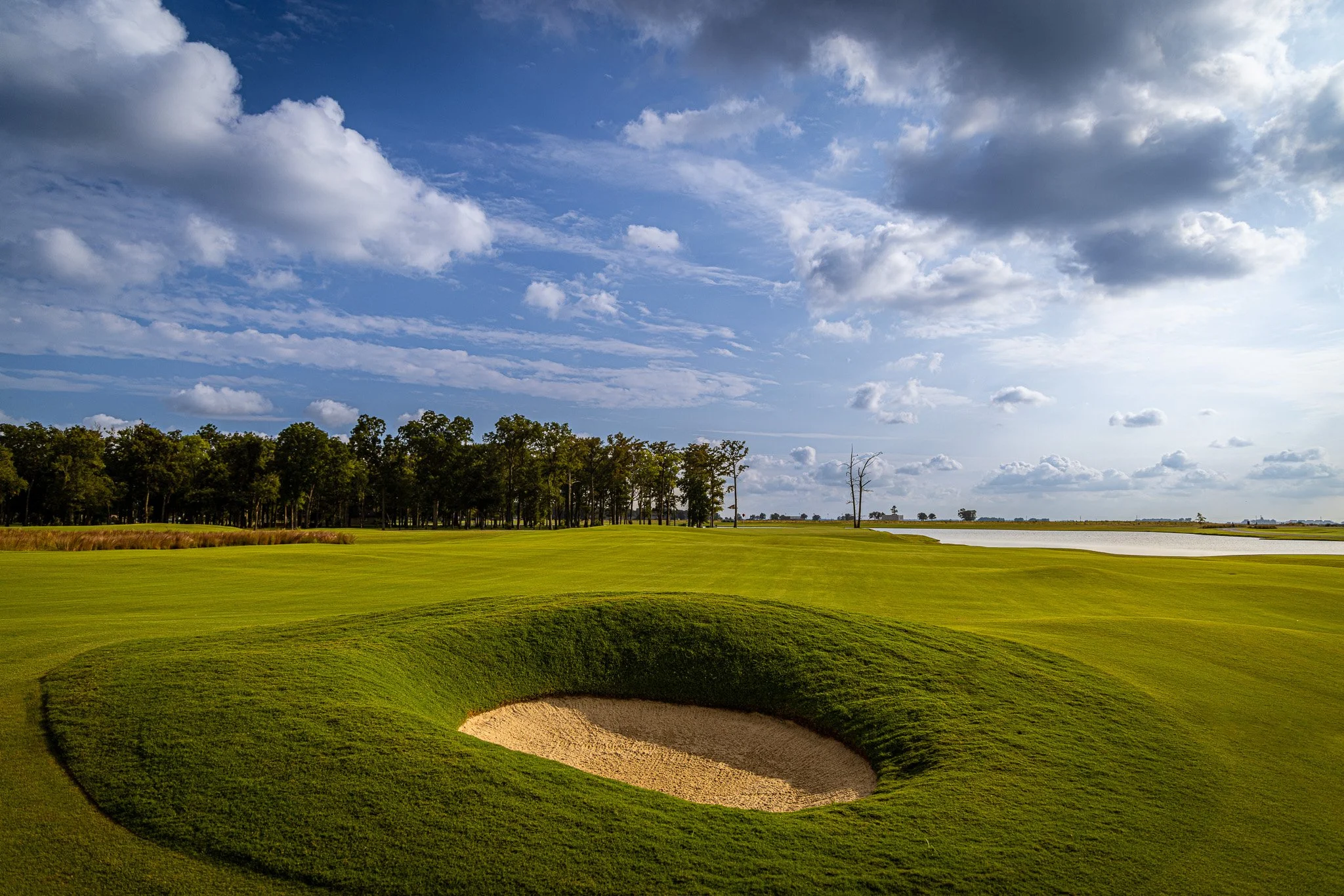 A golf course with a sand bunker in the foreground, green grass, trees in the middle ground, and a partly cloudy sky with sunlight in the background.