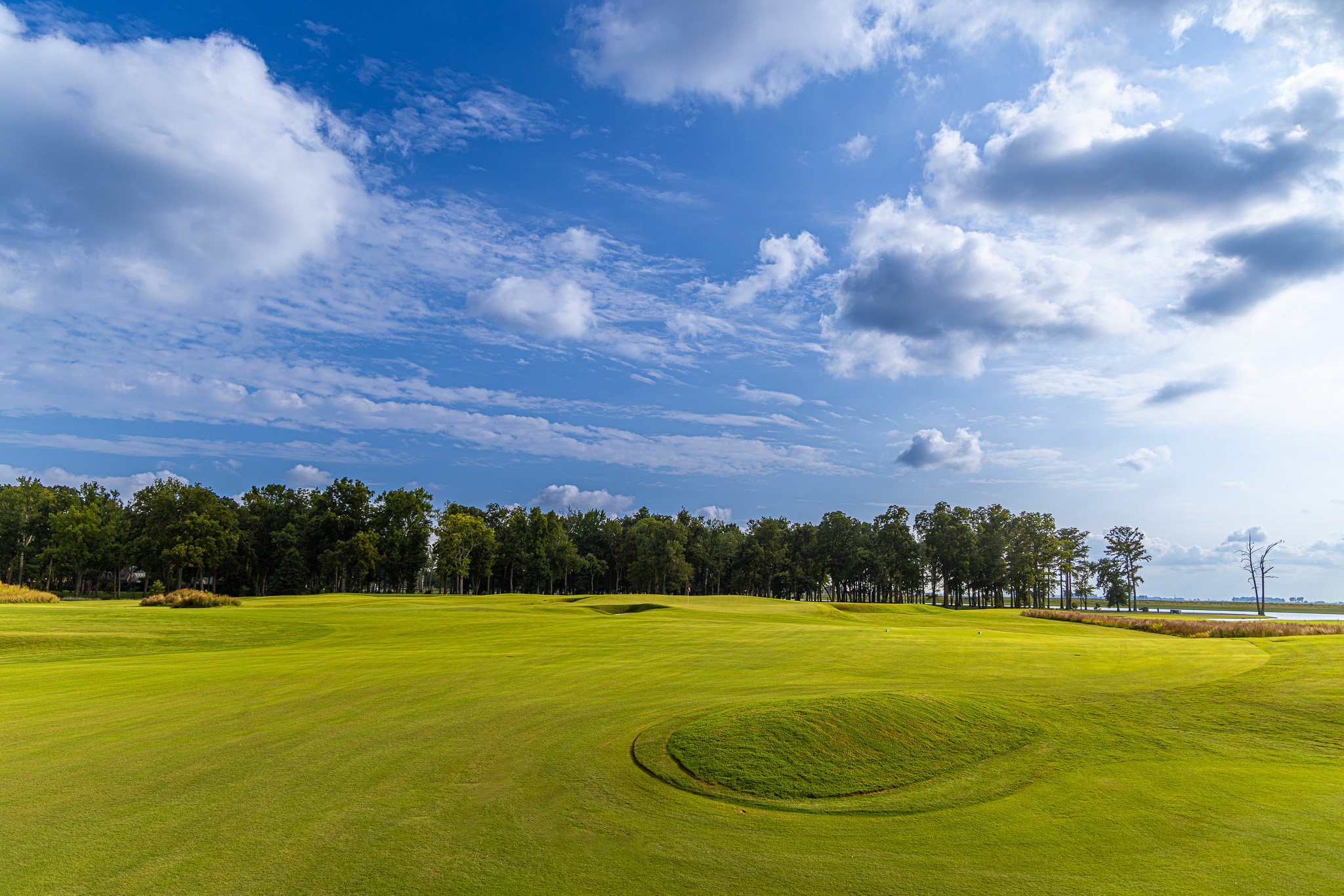 A golf course with neatly trimmed grass, a small sand trap, on a sunny day with blue sky and scattered white clouds, and a line of trees in the distance.