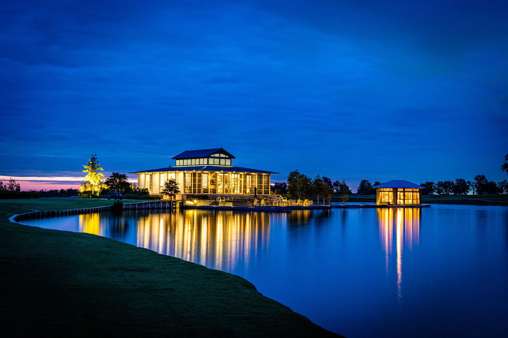 Night scene of lit buildings reflecting on a calm lake with trees and a grassy area in the foreground.