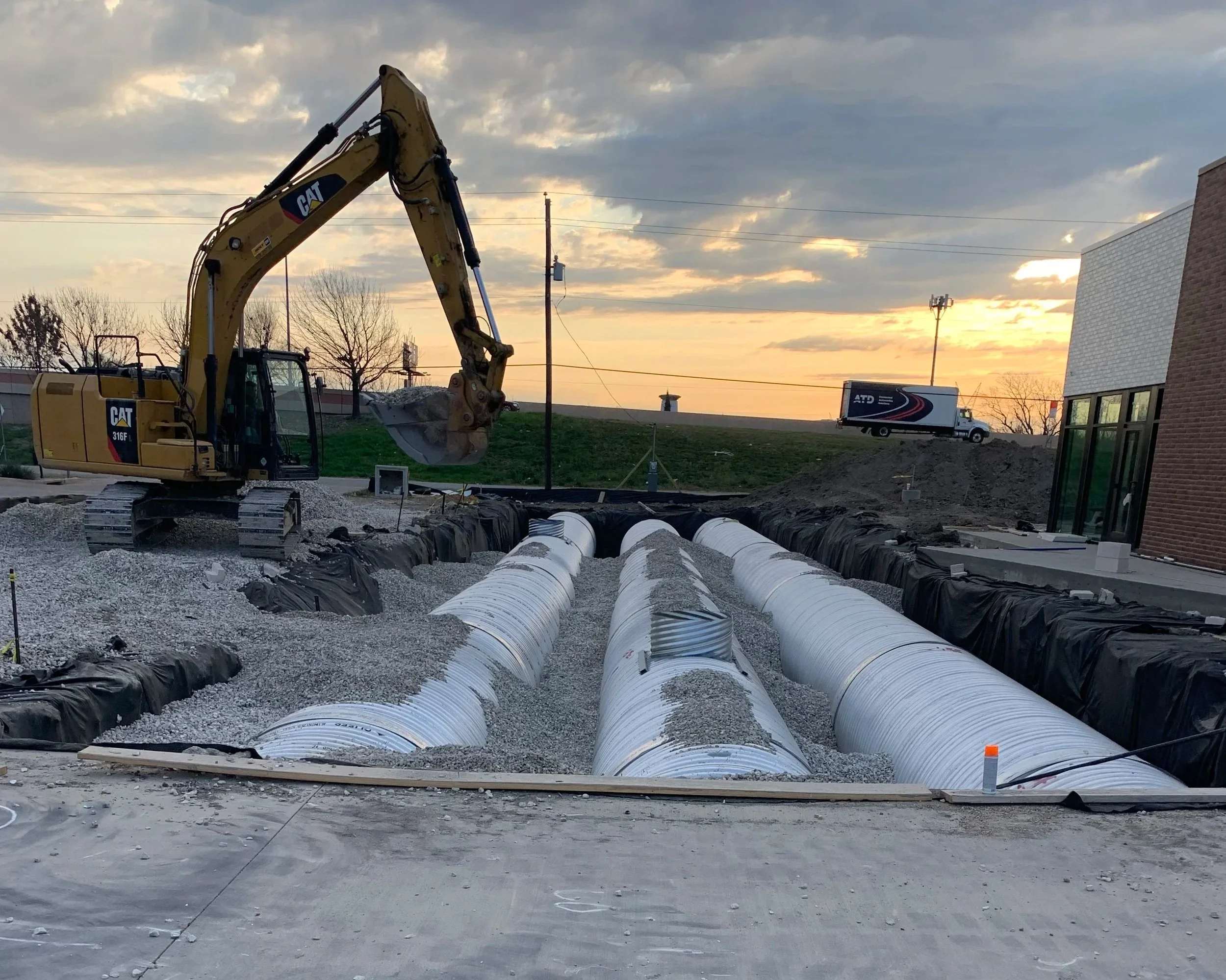 Construction site with a large yellow CAT excavator and several large pipes laid out on the ground, with a building and a truck in the background, during sunset with a partly cloudy sky.