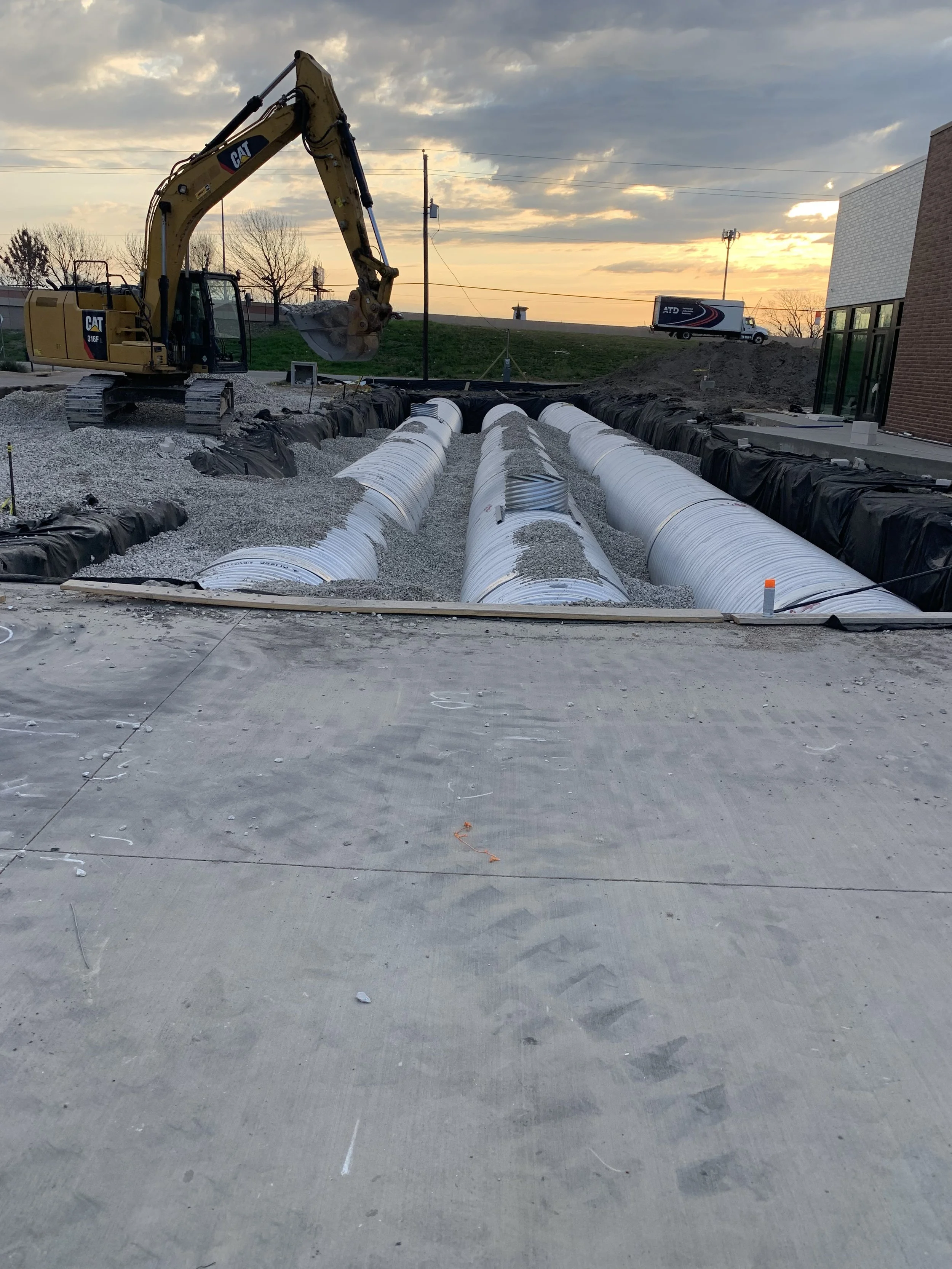 Construction site with large pipes, gravel, and a Caterpillar excavator at sunset.