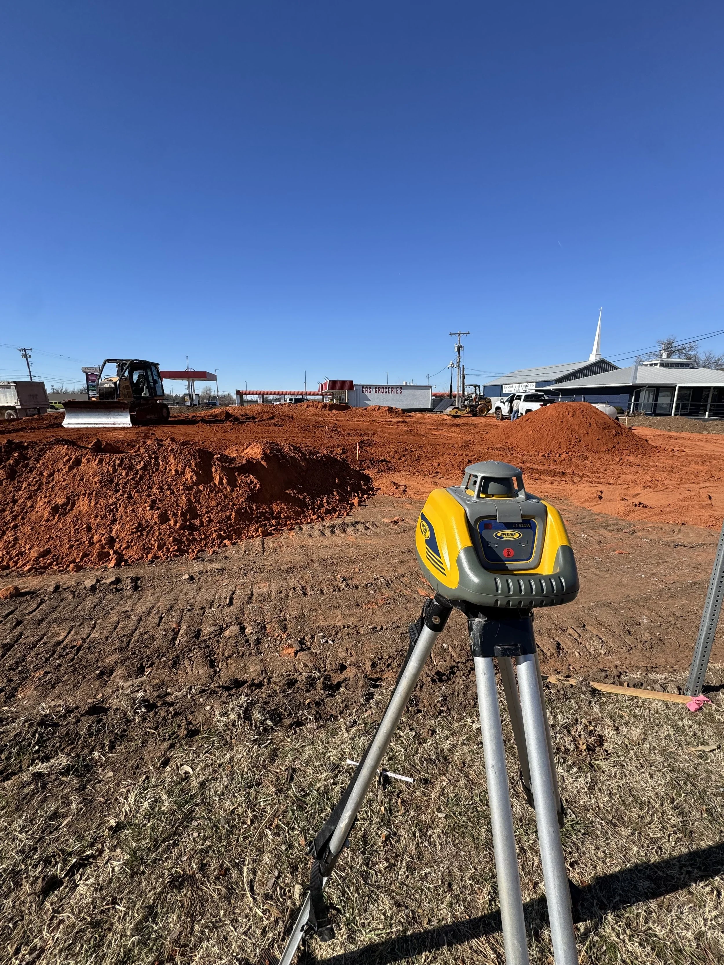 Construction site with a yellow laser level on tripod, dirt piles, machinery, and a gas station in the background under a clear blue sky.