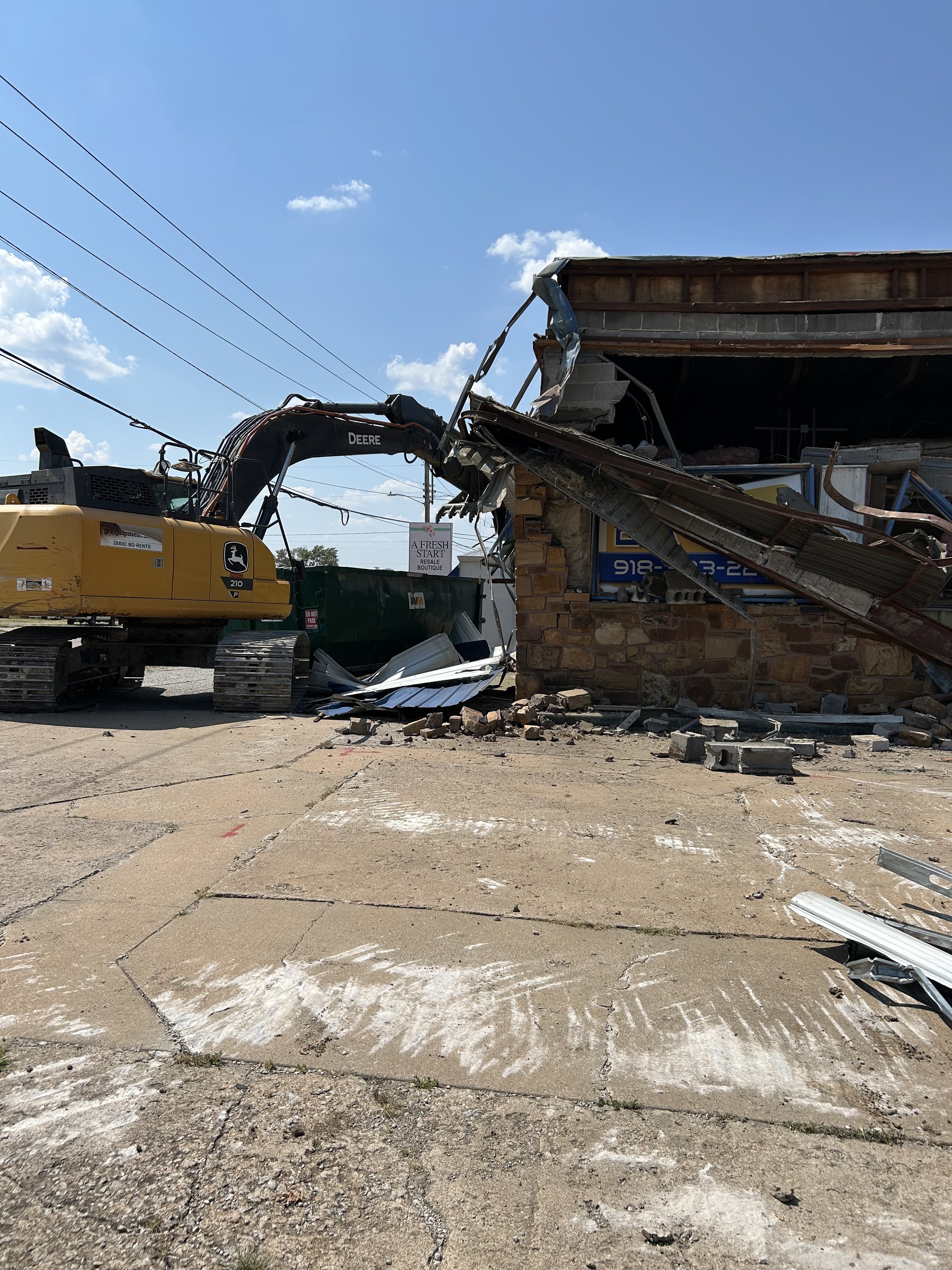 A construction scene with a yellow excavator demolishing part of a building's roof and wall, debris scattered on the ground under a blue sky with a few clouds.