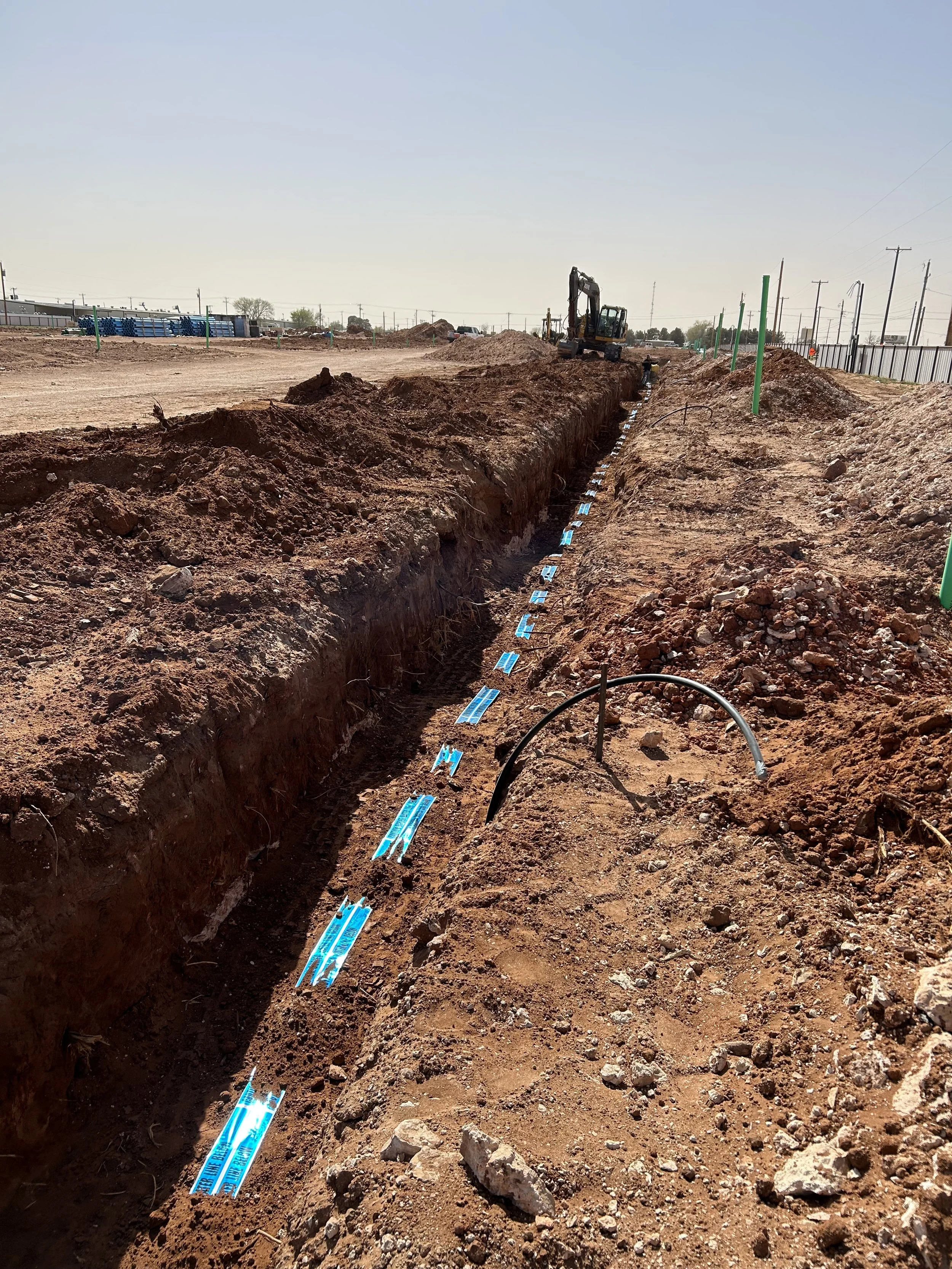 Construction site with a trench, blue markings on the ground, and an excavator in the background.